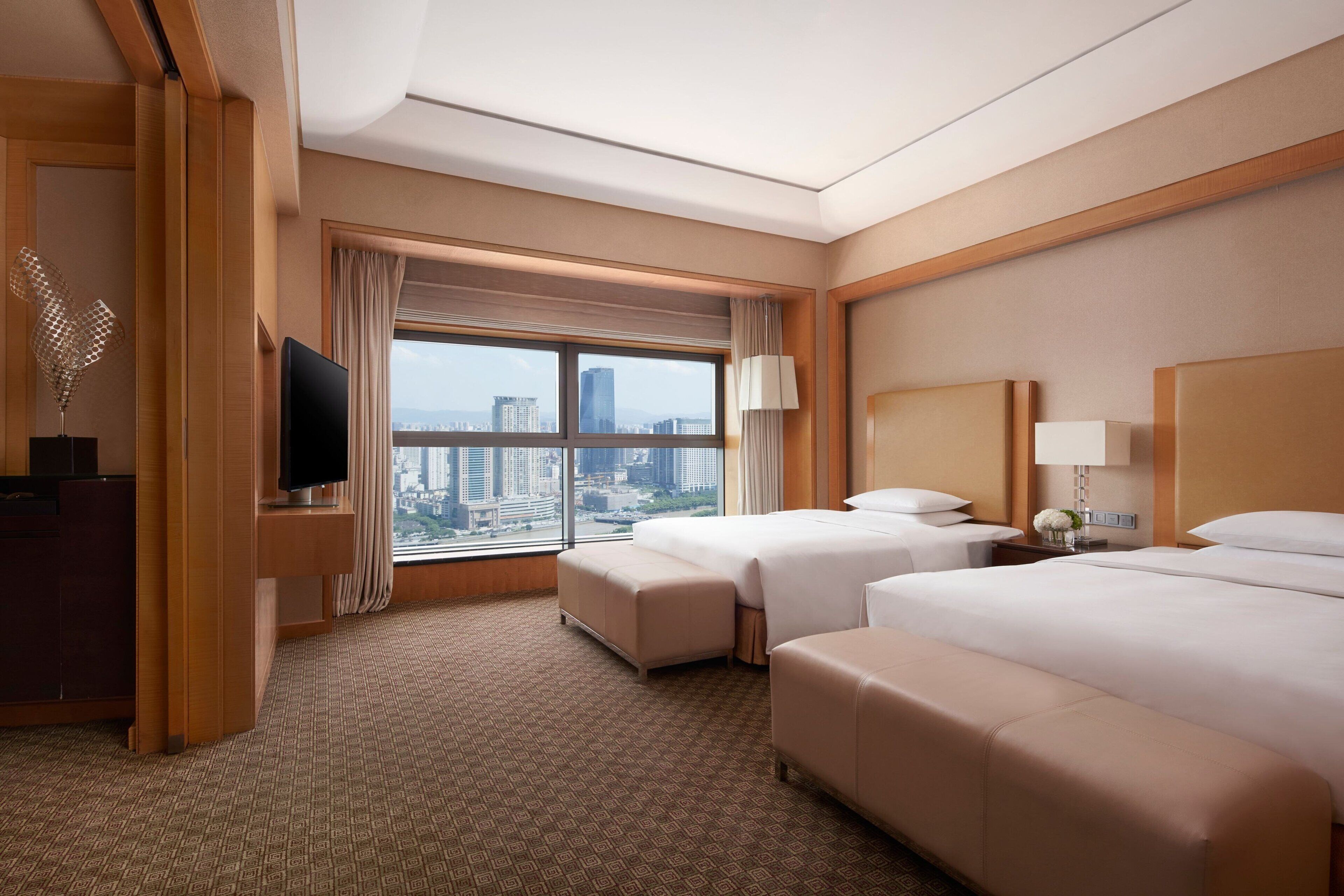Habitación de hotel con dos camas, televisión, escritorio y un gran ventanal con vistas a la ciudad.