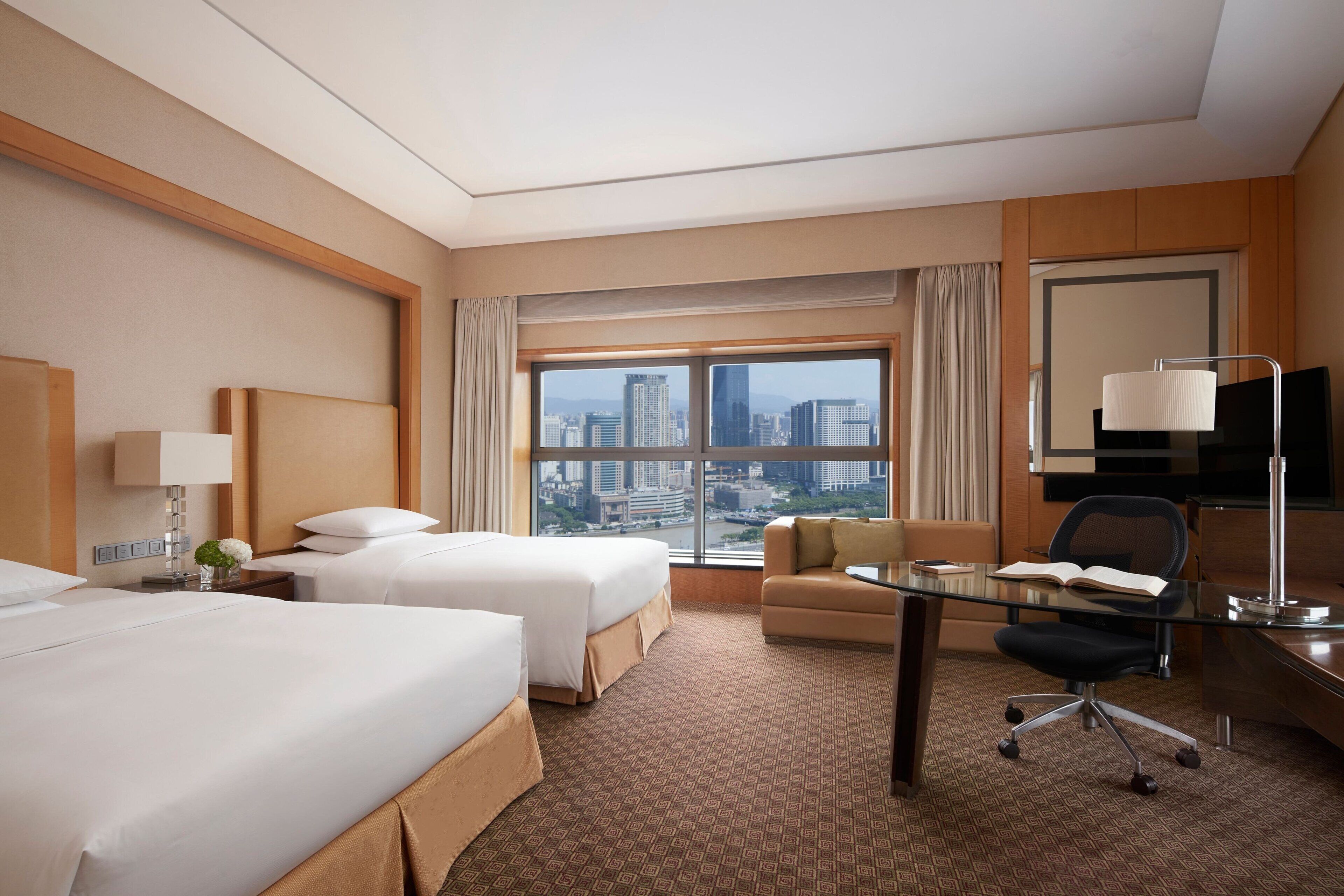 A hotel room with two beds, a sofa, a desk, and a large window offering a city view.