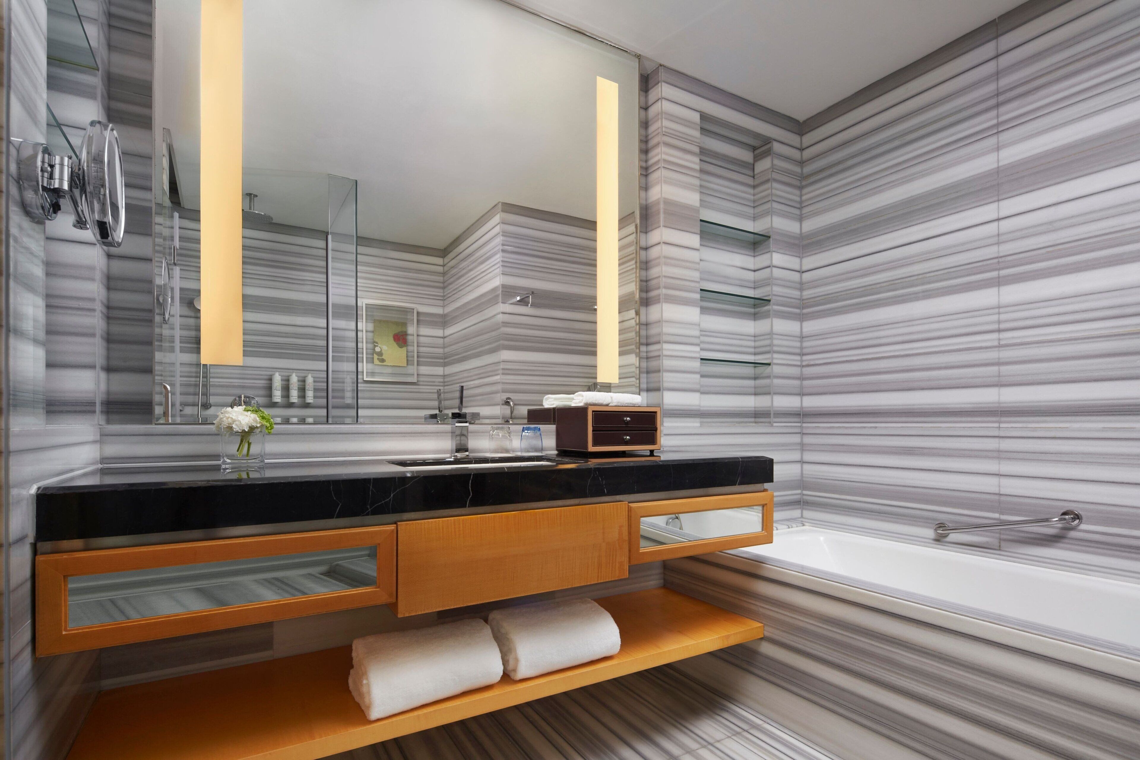 A modern bathroom with a marble backsplash, a black countertop, a wooden vanity, a glass shelf, a white bathtub, and a large mirror.