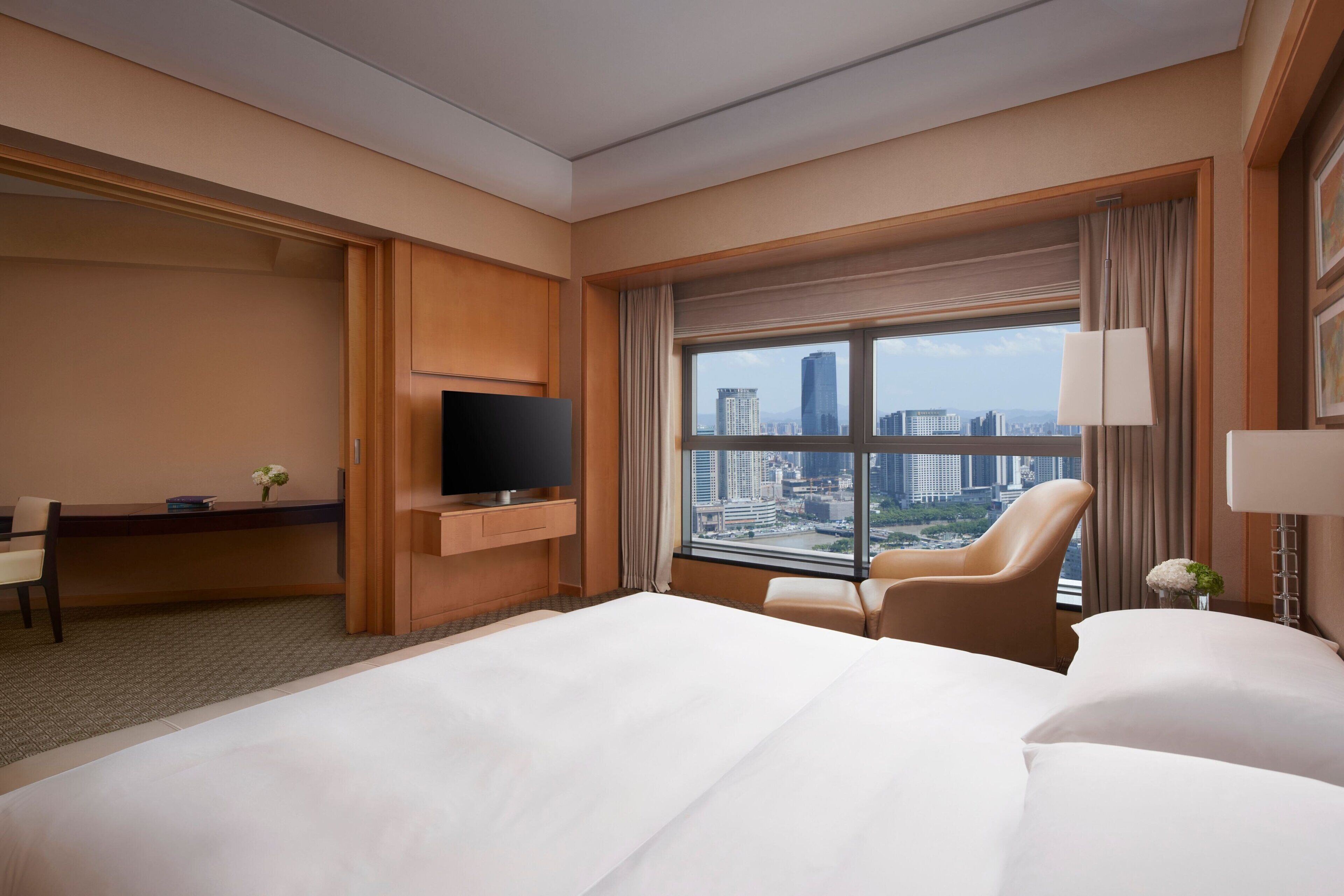 Habitación de hotel con una cama grande, un escritorio con televisor, una silla y una ventana con vistas a la ciudad.