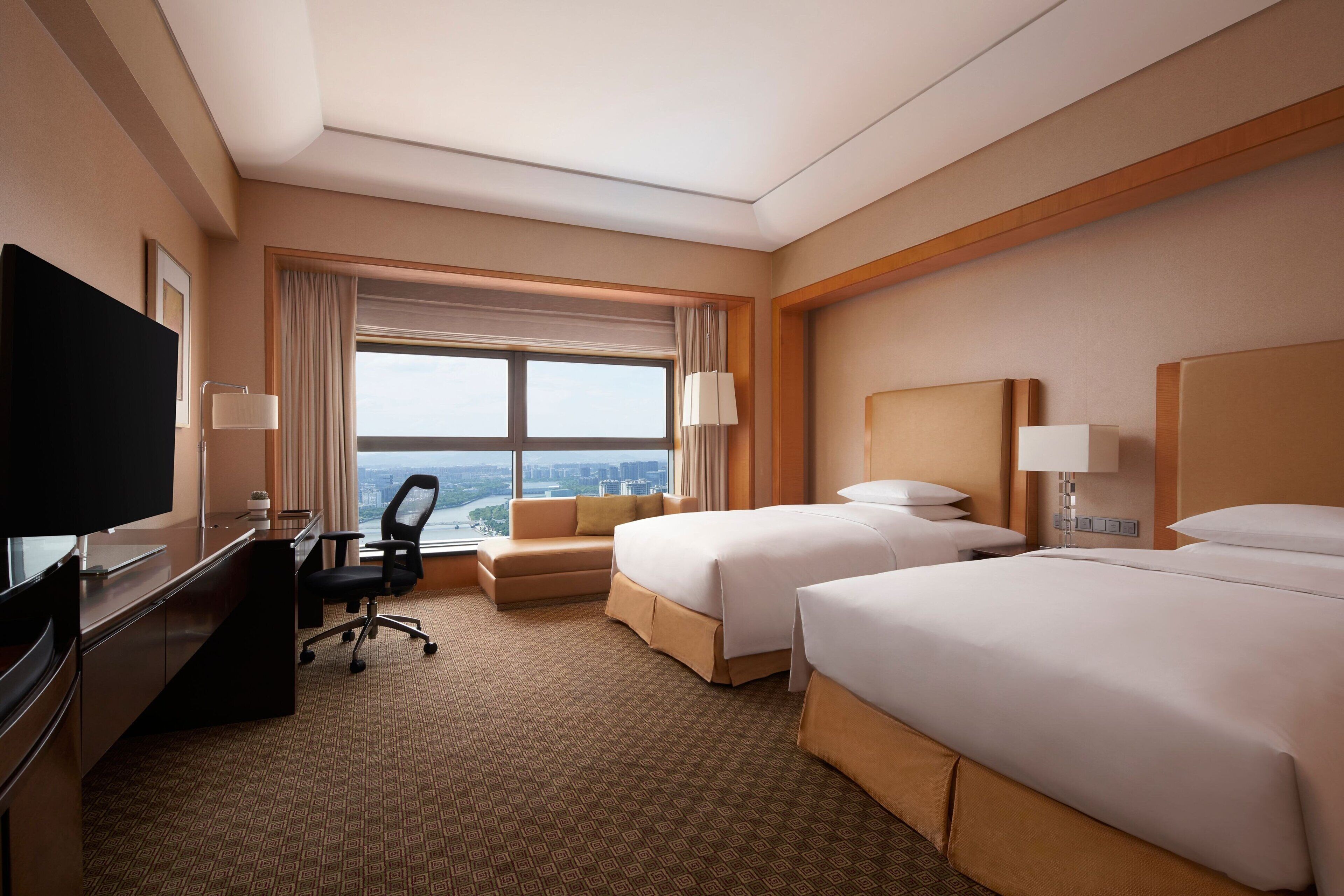 A hotel room with two beds, a desk with a TV, a sofa, and a large window offering a city view.
