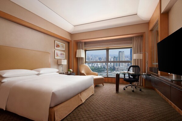 A hotel room with a large bed, a desk with a chair, a television, and a view of a cityscape through a large window.