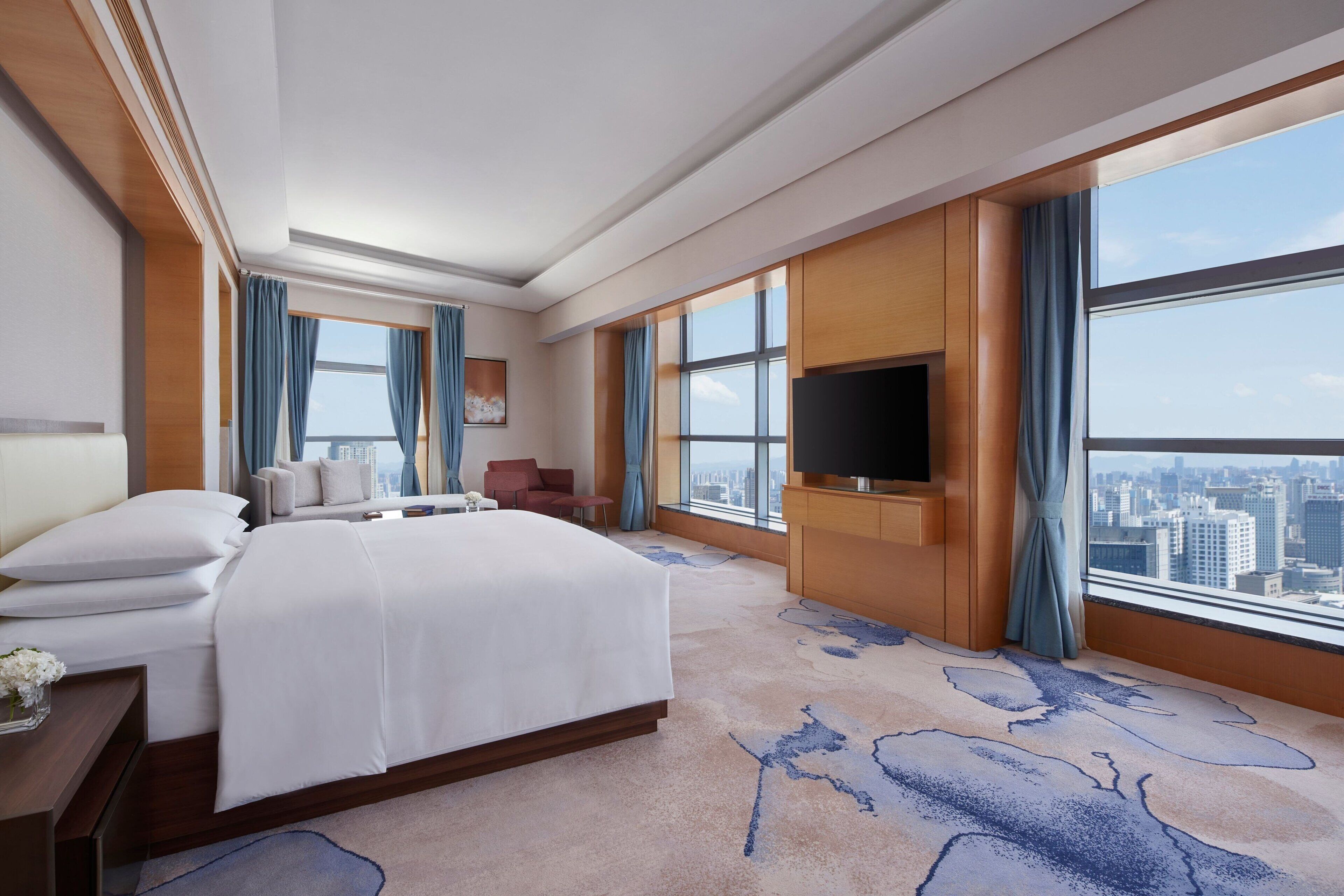 A hotel room with a large bed, a flat-screen TV mounted on the wall, a city view through large windows, and a carpet with a blue abstract design.