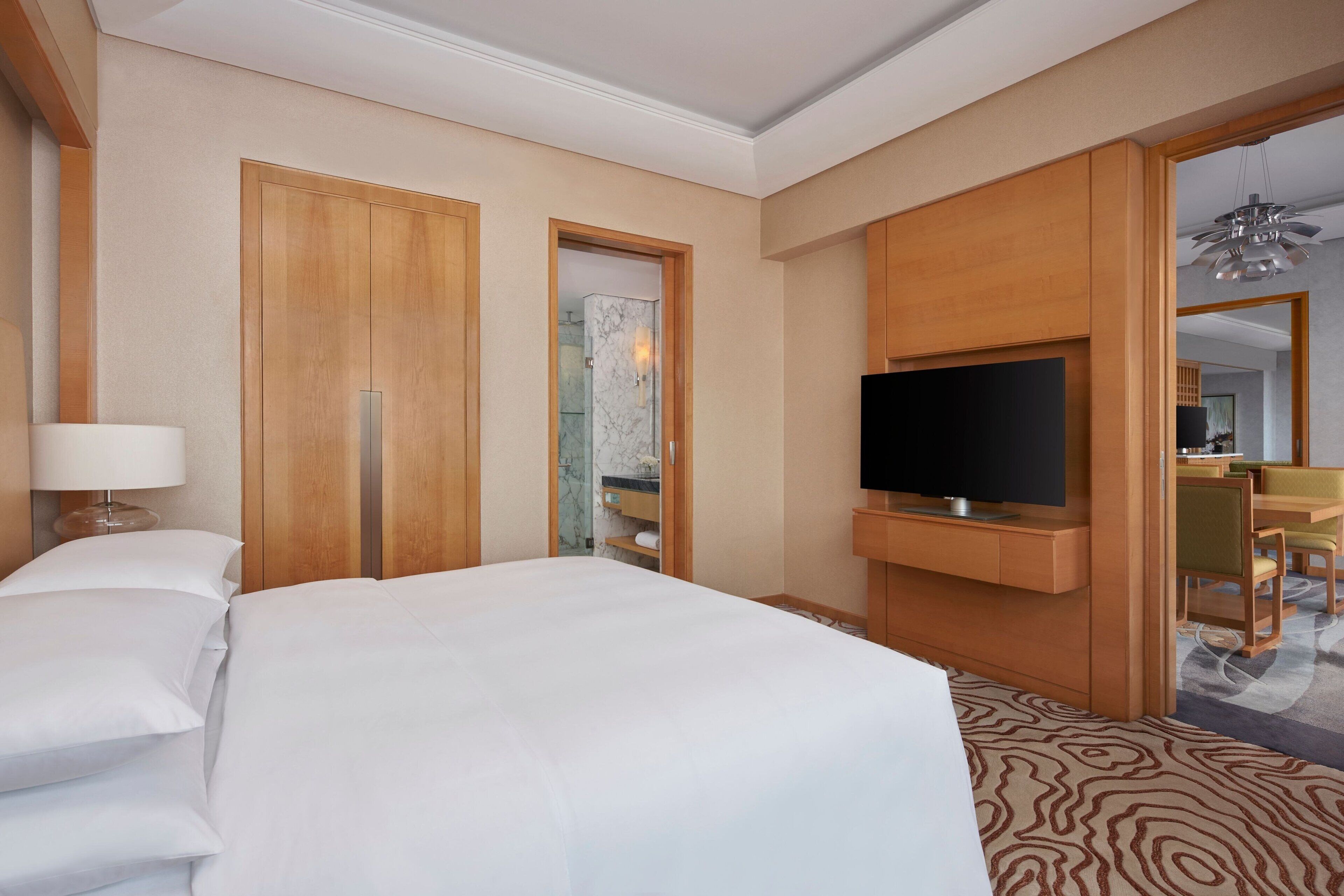 A hotel room with a large bed, a TV mounted on the wall, wooden wardrobes, and a bathroom visible through an open door.