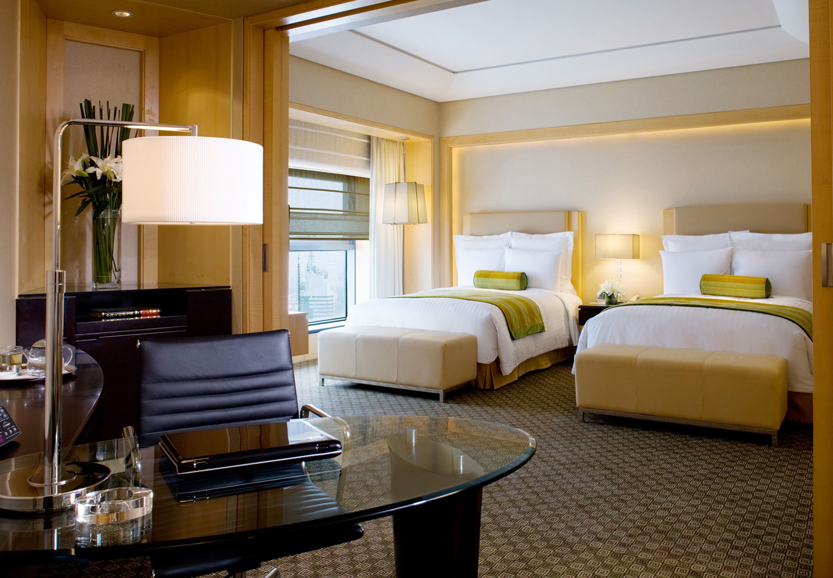 A hotel room with a glass table, two beds with green and white bedding, a desk with a lamp, and a vase with flowers.