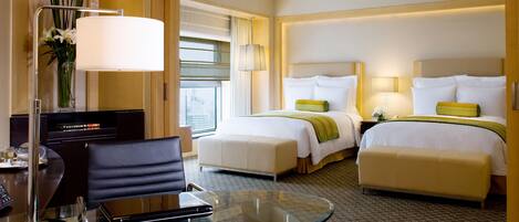 A hotel room with a glass table, two beds with green and white bedding, a desk with a lamp, and a vase with flowers.