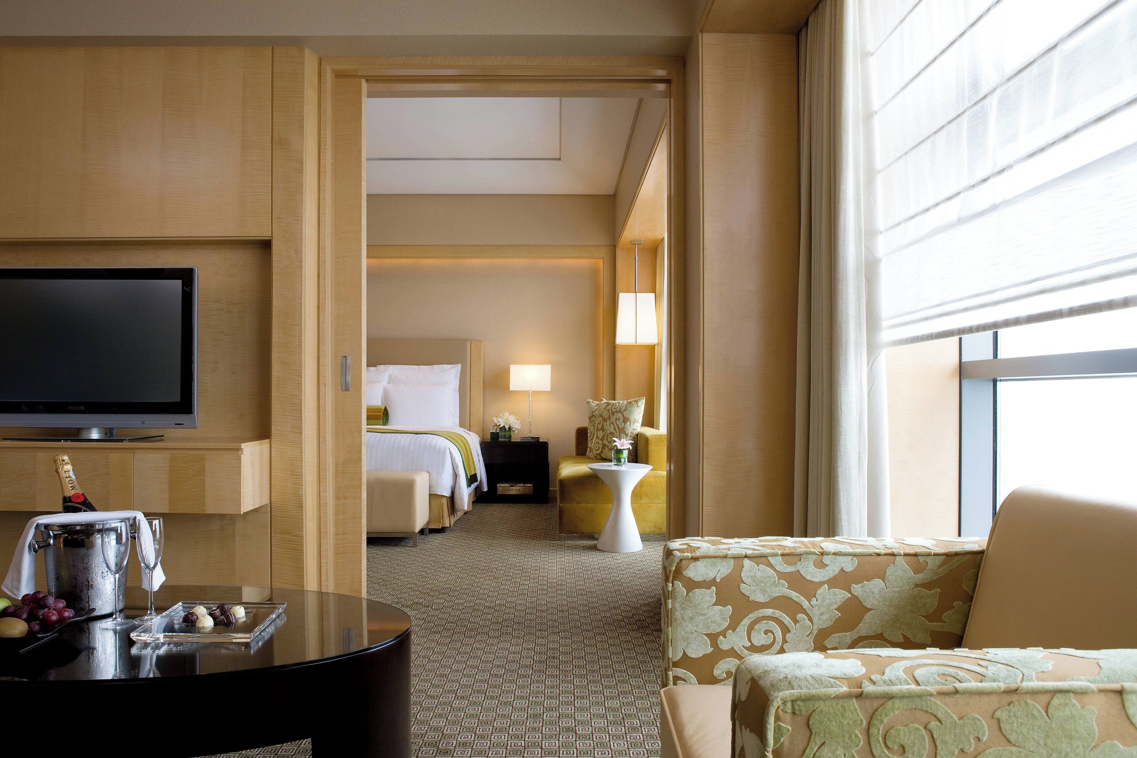 A hotel room with a TV, a minibar, a bed, a sitting area with a chair and a table, and a large window.