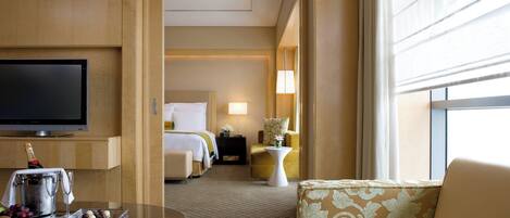 A hotel room with a TV, a minibar, a bed, a sitting area with a chair and a table, and a large window.