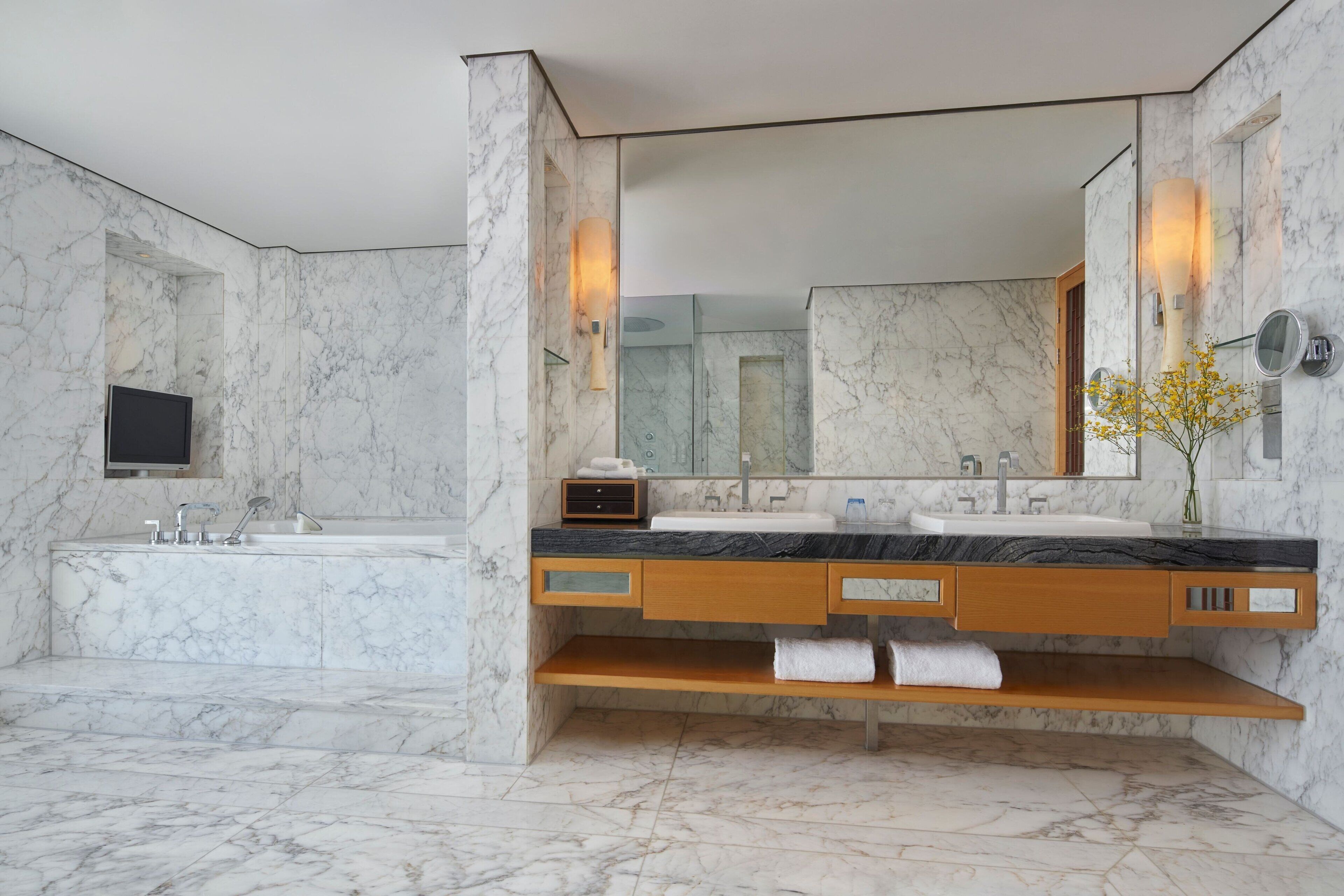 Un baño moderno con suelo de mármol, un lavabo grande, un espejo y un televisor instalado en la pared.