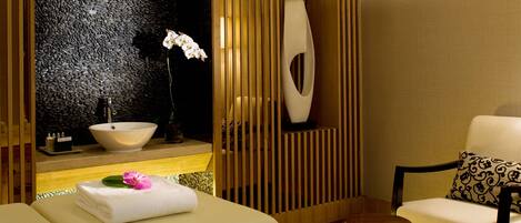 Un moderno spa con una bañera grande, un panel de pared de madera, un lavabo blanco y una silla con un cojín estampado.