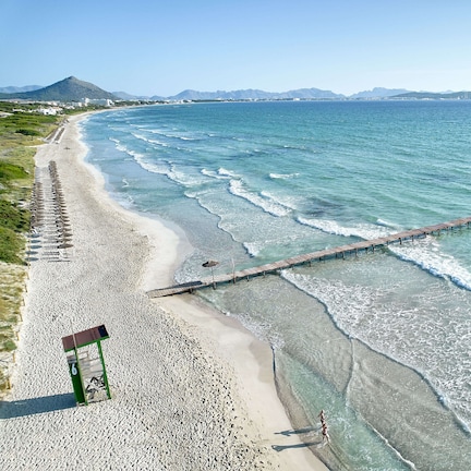 A beach with a wooden pier extending into the water, a lifeguard tower, and mountains in the background.