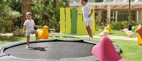 A trampoline in a grassy area with a pool and a building in the background.