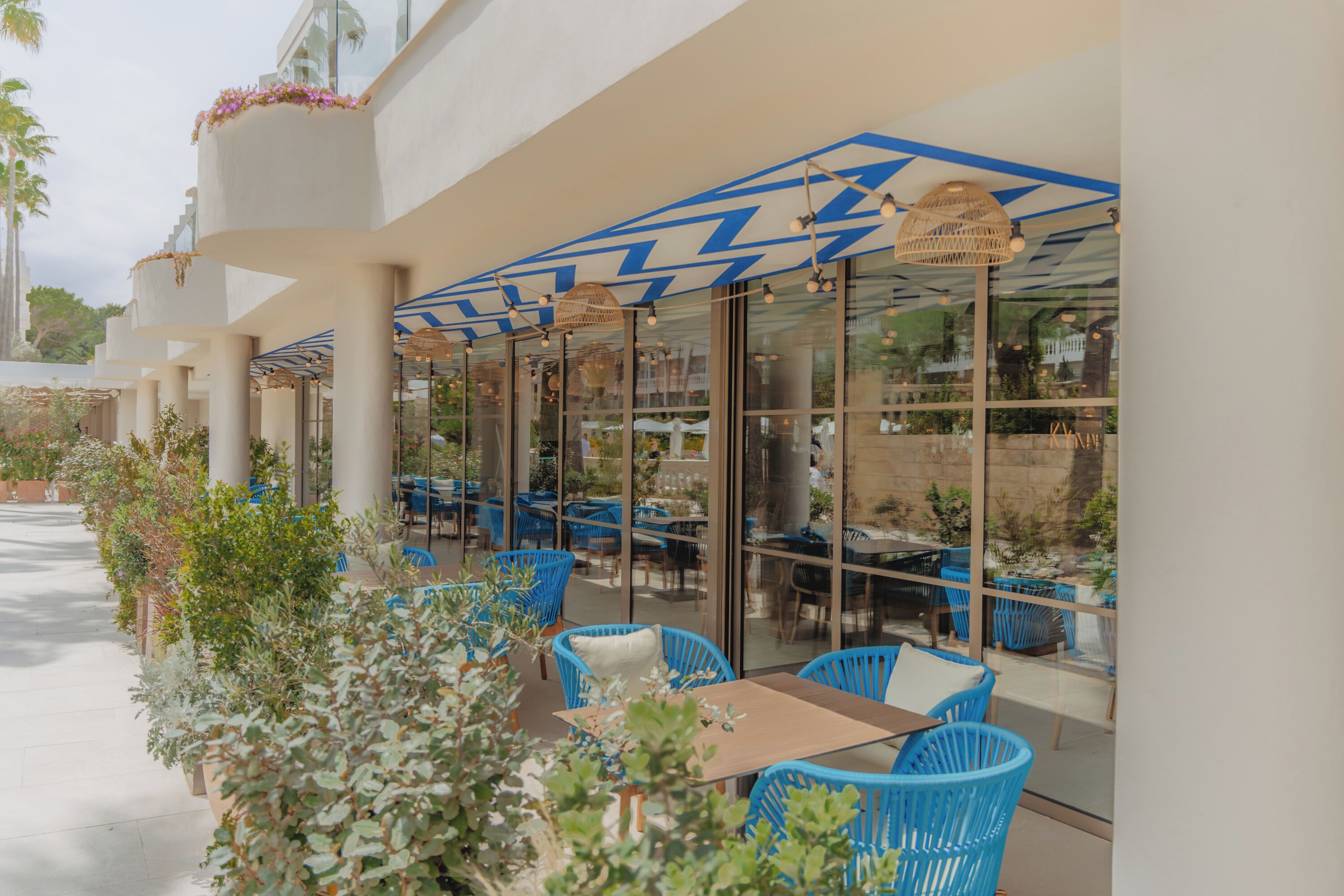 Un área de café al aire libre con sillas y mesas azules, rodeada de paredes de vidrio y plantas en macetas.