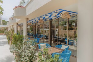 Un área de café al aire libre con sillas y mesas azules, rodeada de paredes de vidrio y plantas en macetas.