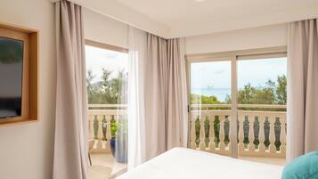A hotel room with a bed, a TV mounted on the wall, and a balcony with a view of the sea and greenery.