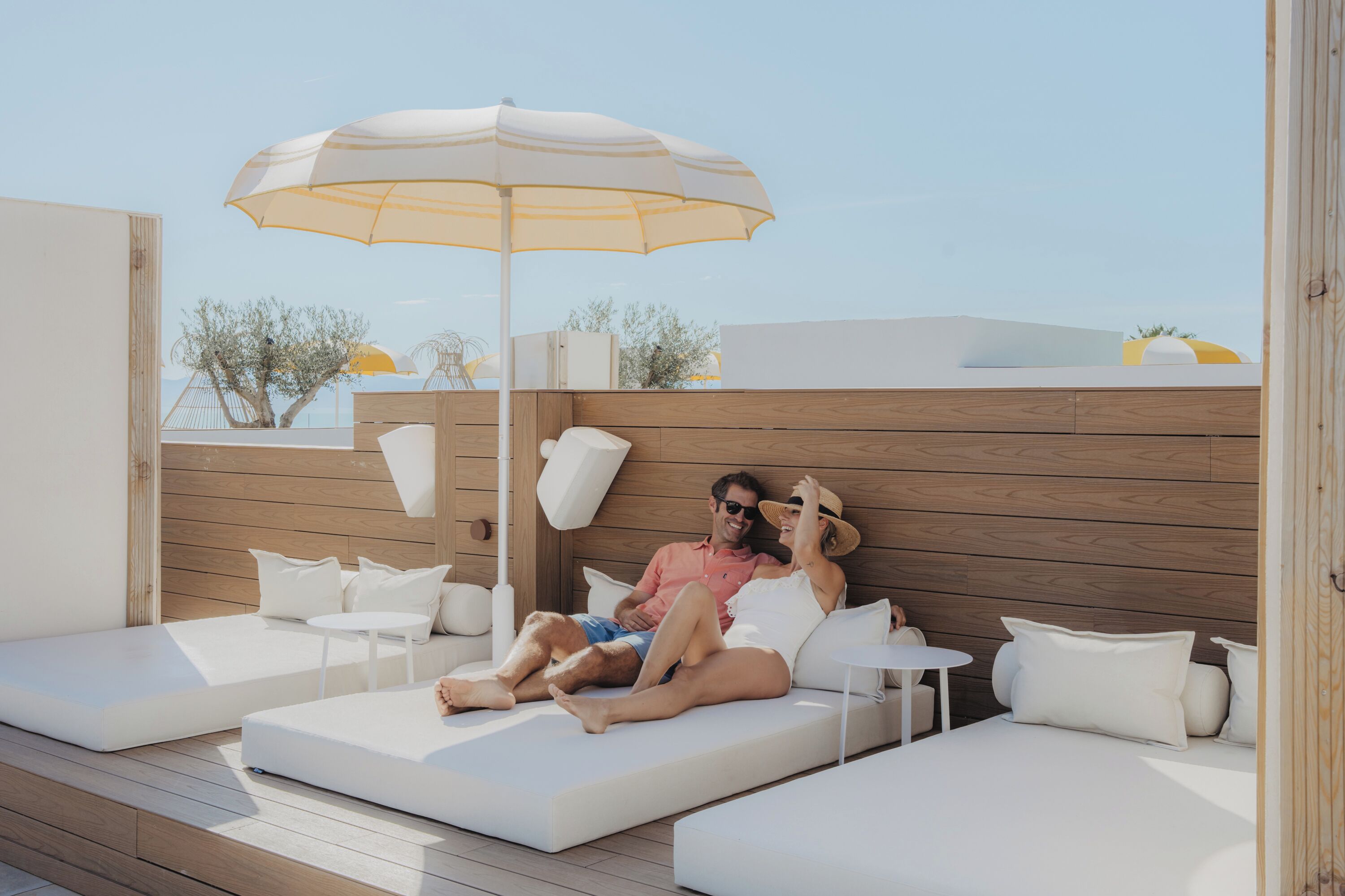 A couple relaxing on a white outdoor sofa with a wooden backdrop, under a large umbrella, with a clear view of the sky and distant landscape.