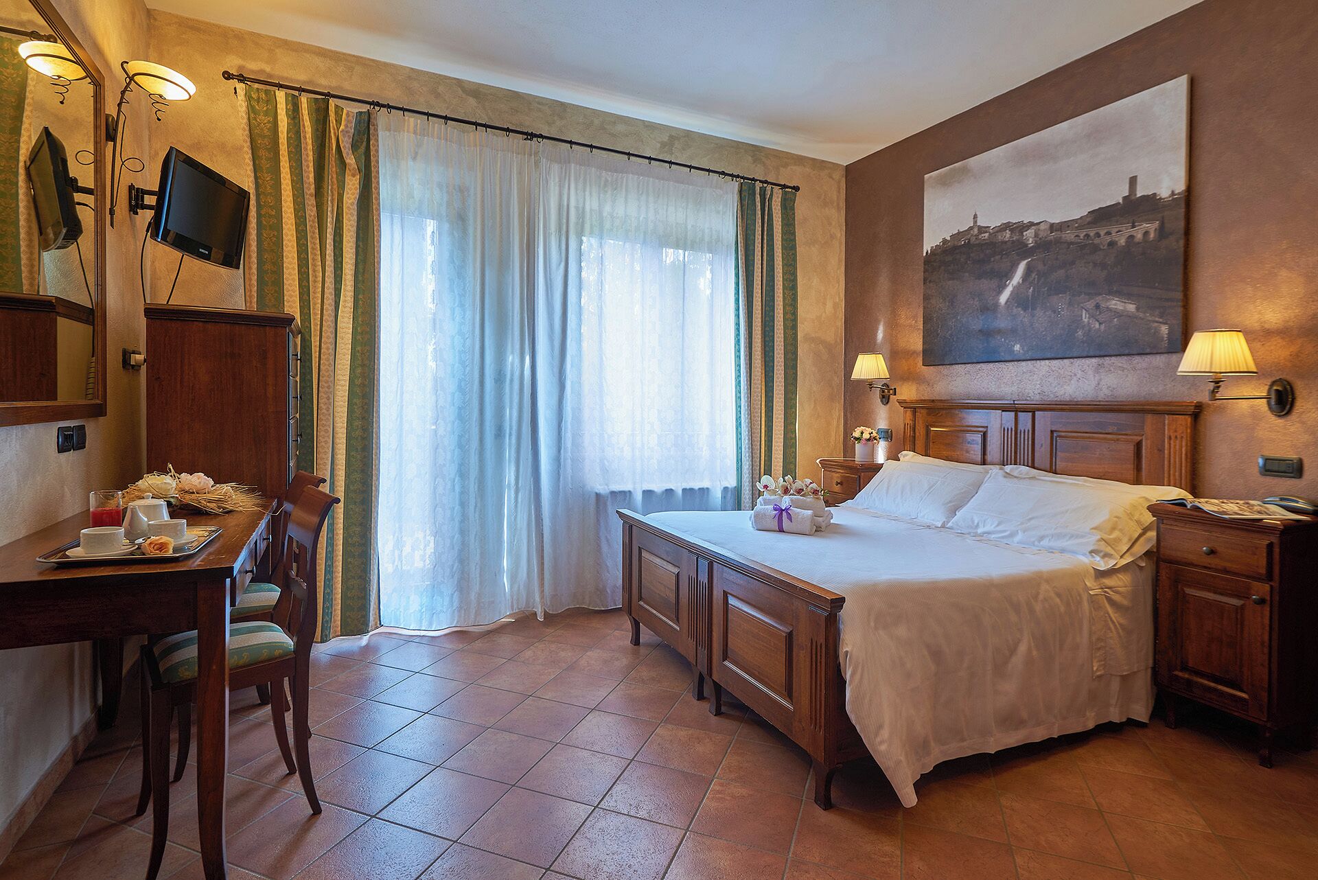 A hotel room with a bed, bedside table, desk, chair, and a painting on the wall.