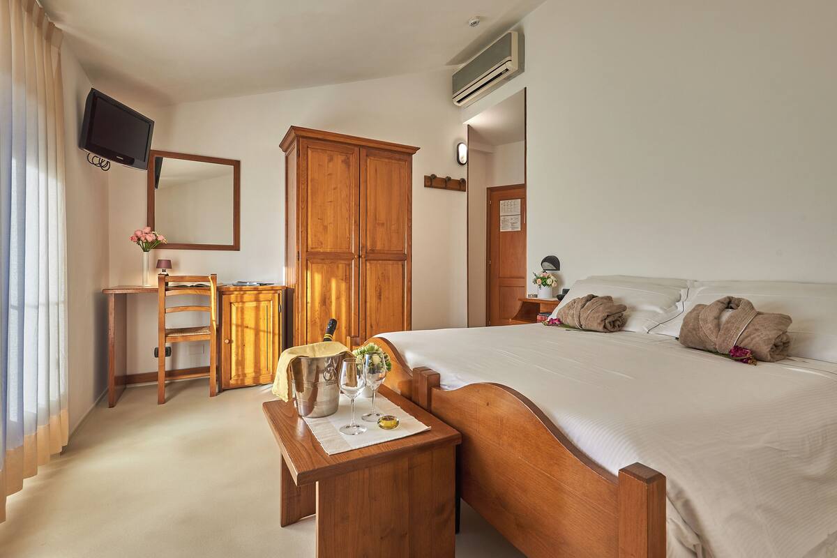 A hotel room with a wooden bed, a desk with a chair, a large wardrobe, a television mounted on the wall, and an air conditioning unit.