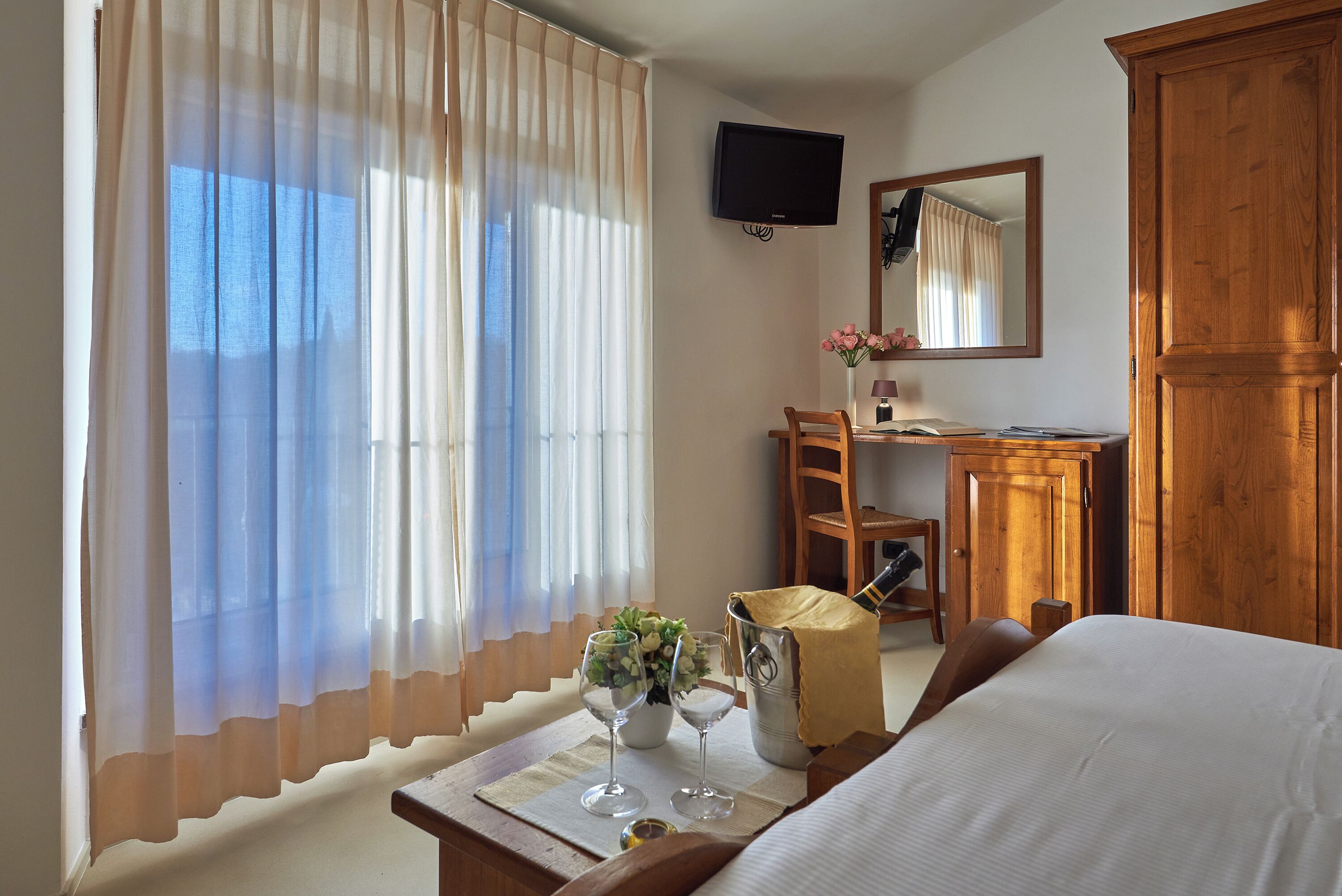 A hotel room with a bed, a desk with a chair, a television, and a large wooden wardrobe.