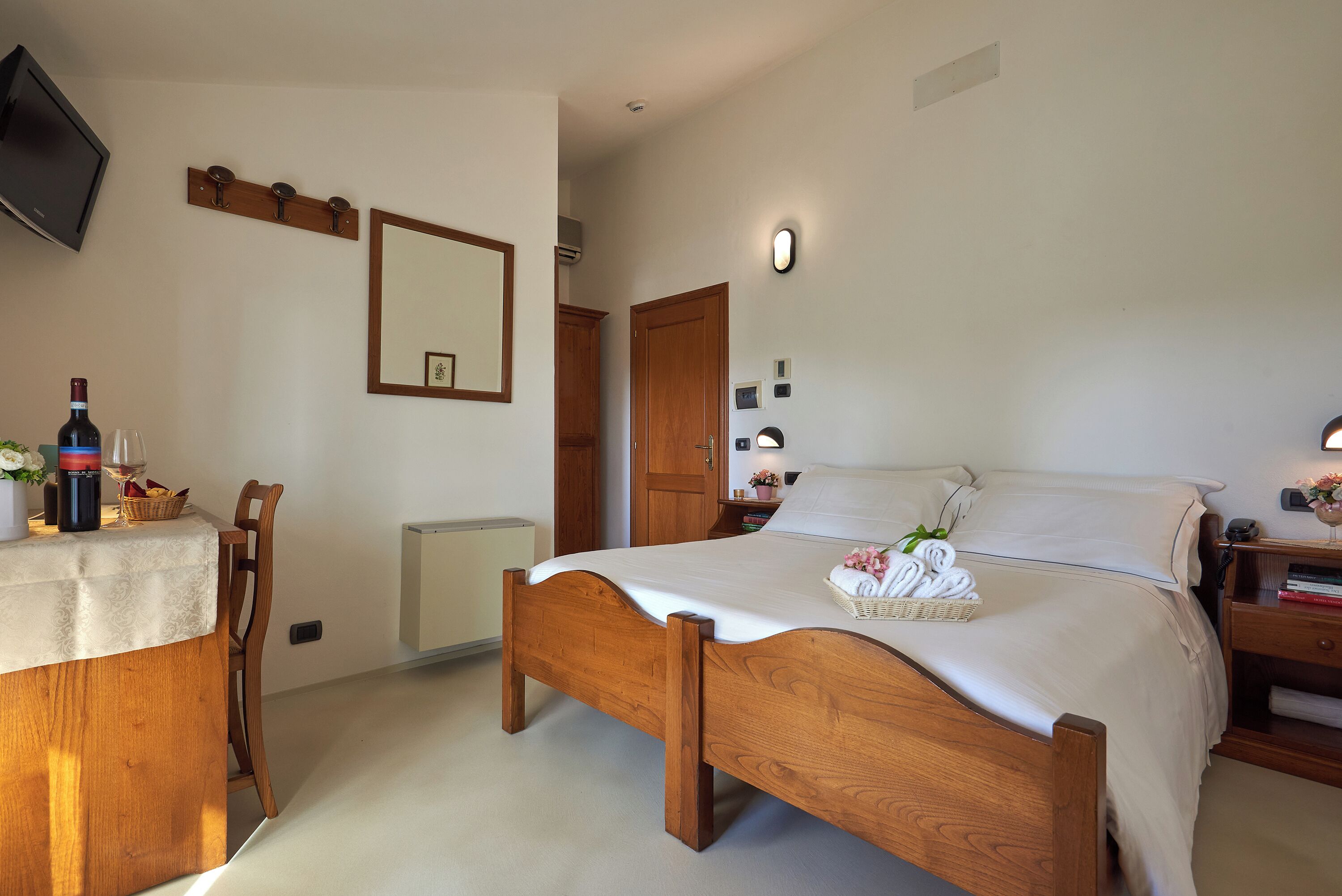 A bedroom with a wooden bed, a bedside table, a chair, a desk, and a television mounted on the wall.