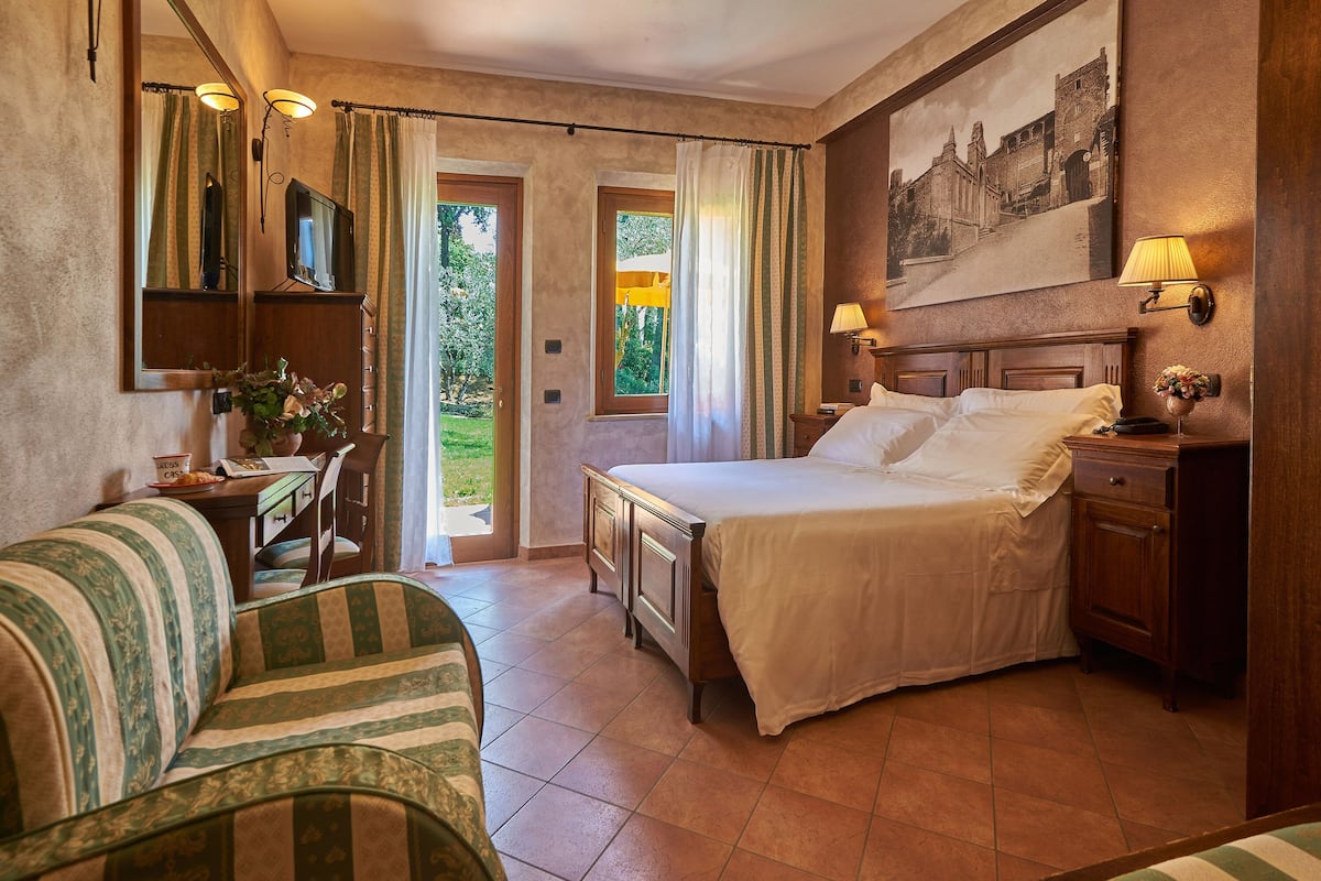 A hotel room with a bed, a sofa, a desk with a chair, a TV, and a painting on the wall.