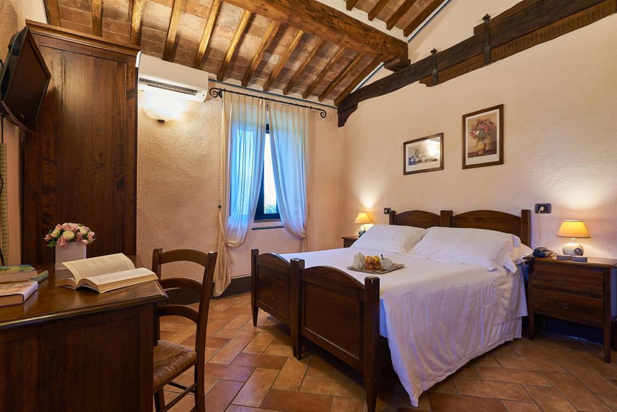 A bedroom with a wooden bed, a desk with a book, a chair, a window with curtains, and a nightstand with a lamp.