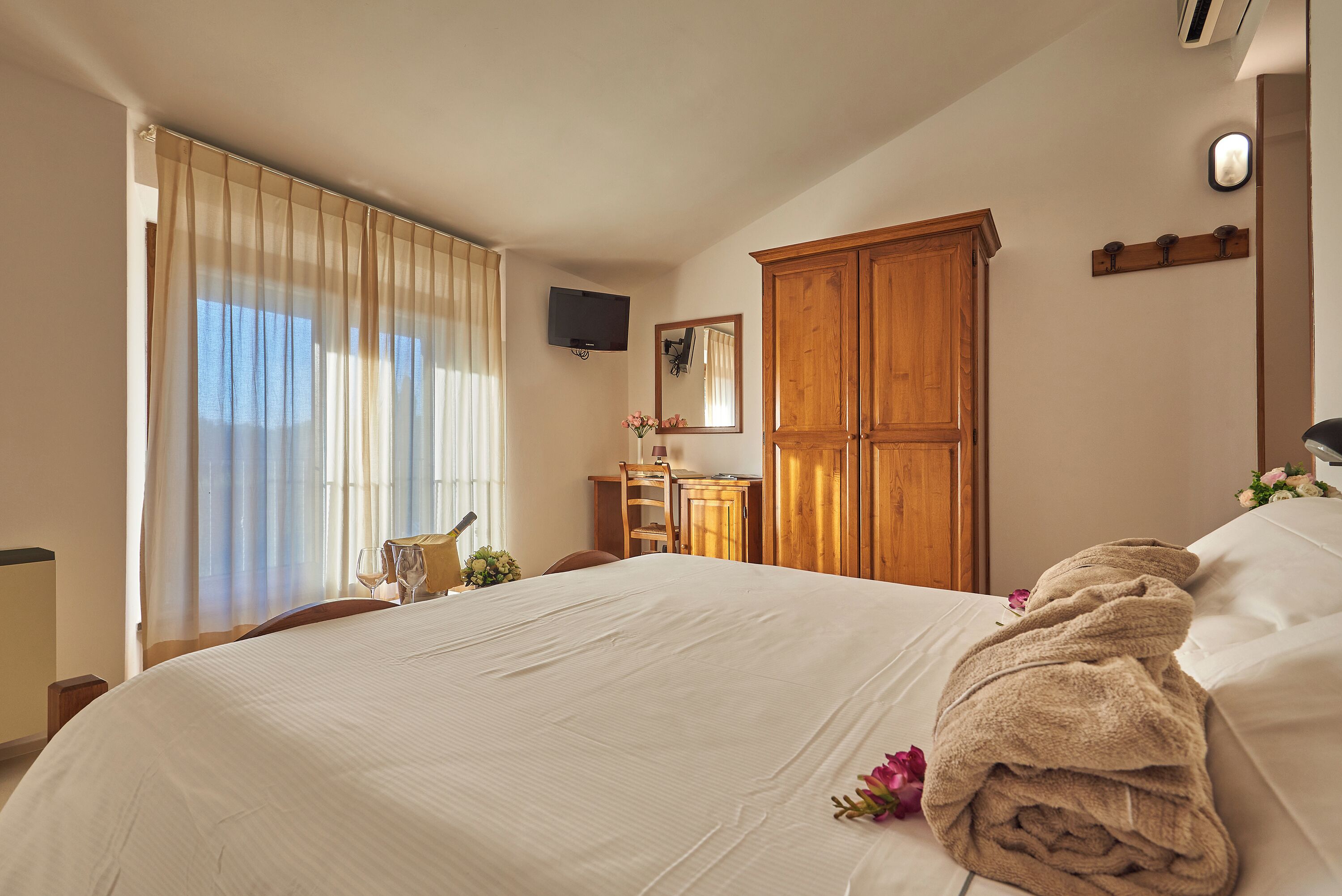 A hotel room with a large bed, a wooden wardrobe, a desk with a chair, and a television mounted on the wall.