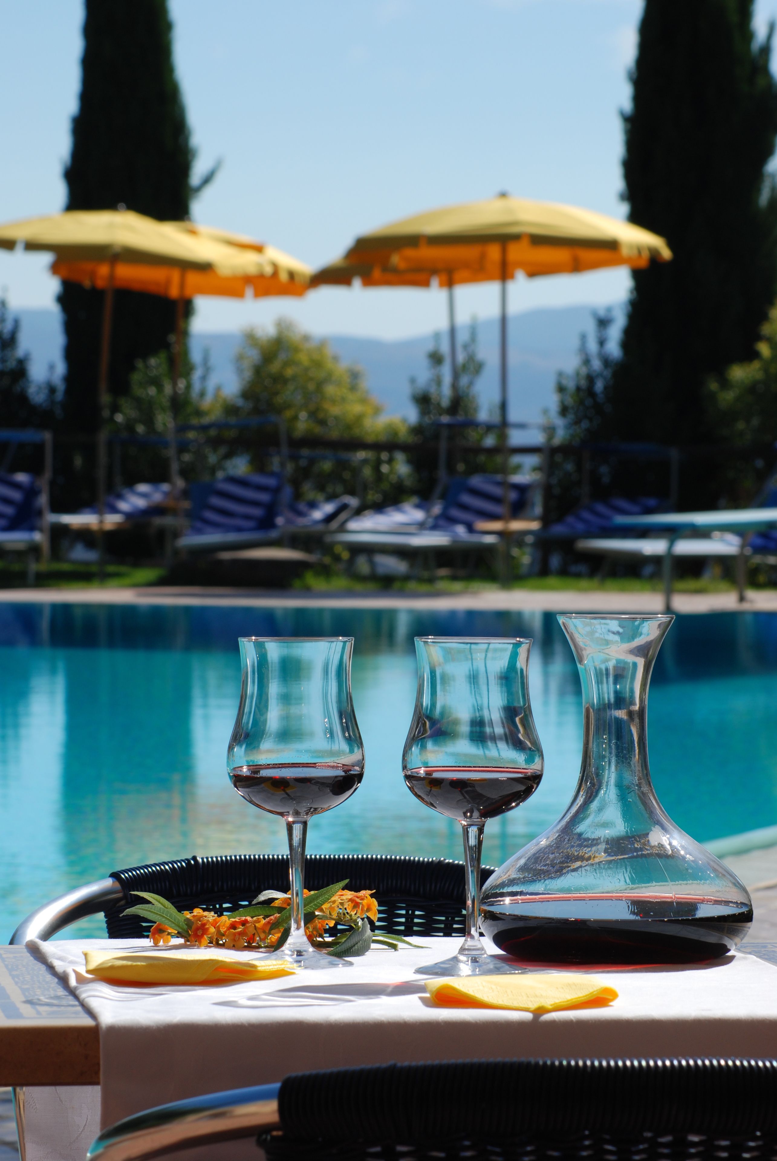 Une table placée au bord de la piscine, avec deux verres à vin et un vase, entourée de chaises longues et de parasols.