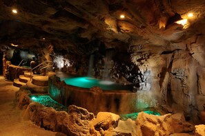 Interior que se asemeja a una cueva, con una piscina termal central, formaciones rocosas y una escalera que conduce a un nivel superior.