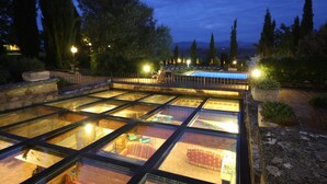 A glass-enclosed pool area with multiple levels and seating, surrounded by trees and stone walls.
