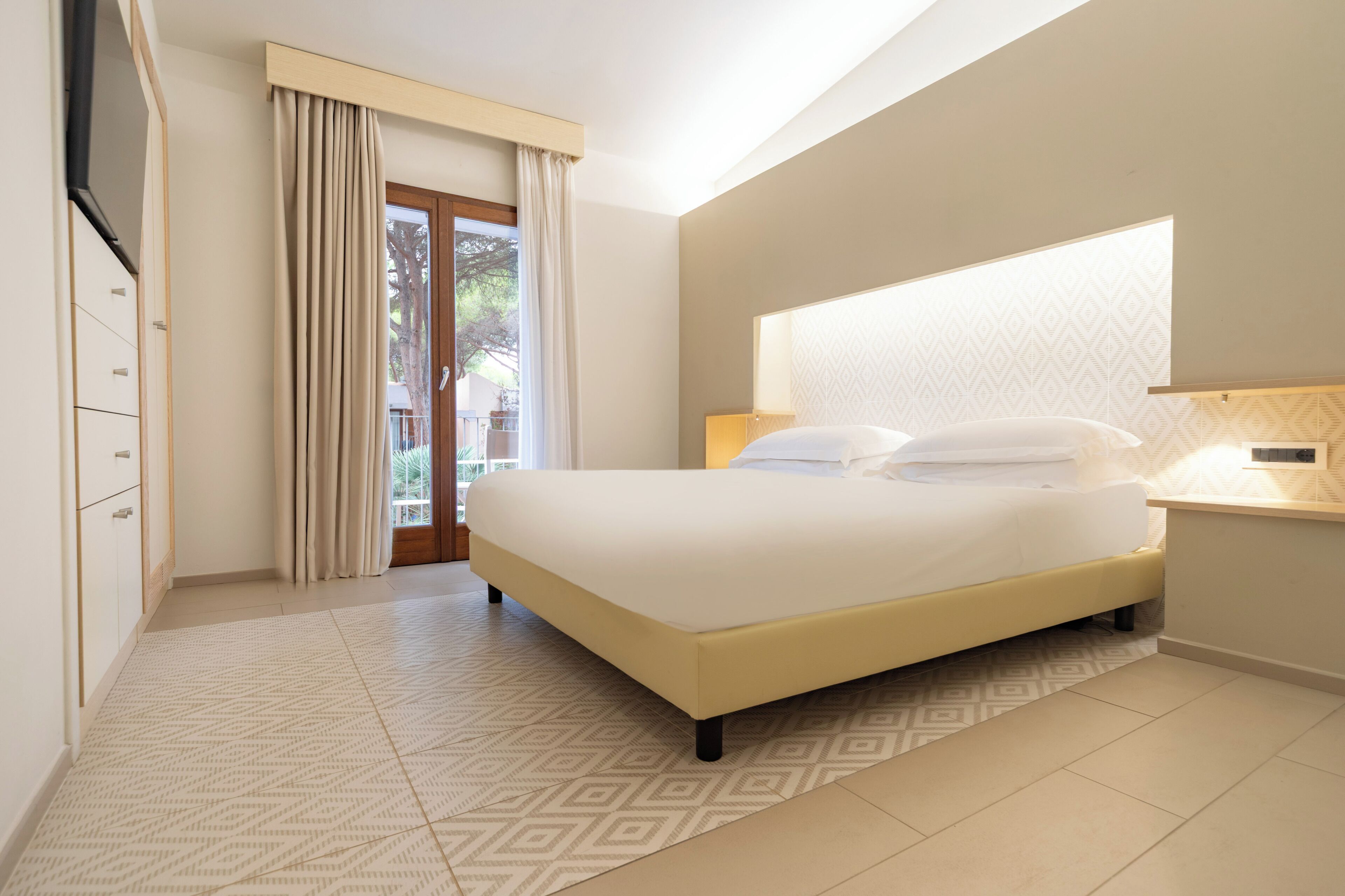 A modern hotel room with a large bed, a TV mounted on the wall, and a view of the outdoors through a glass door.