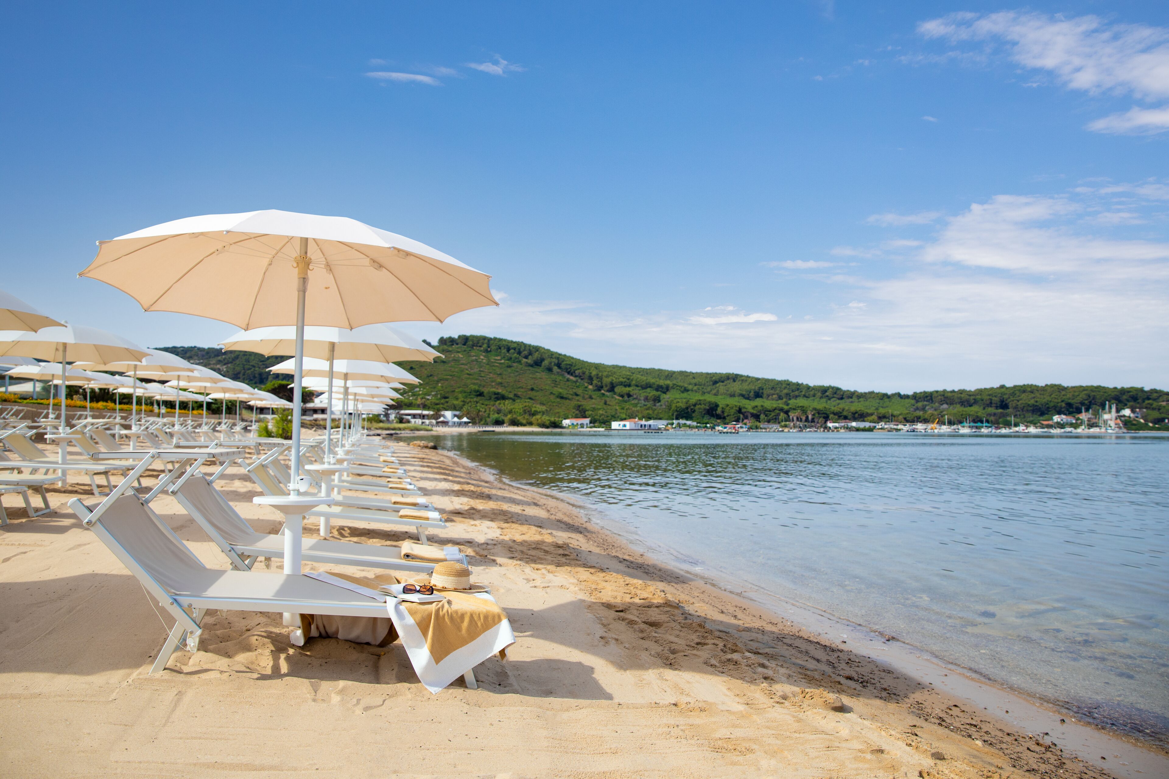 Una spiaggia con file di lettini bianchi e ombrelloni color beige, un lago calmo e un paesaggio collinare sullo sfondo.