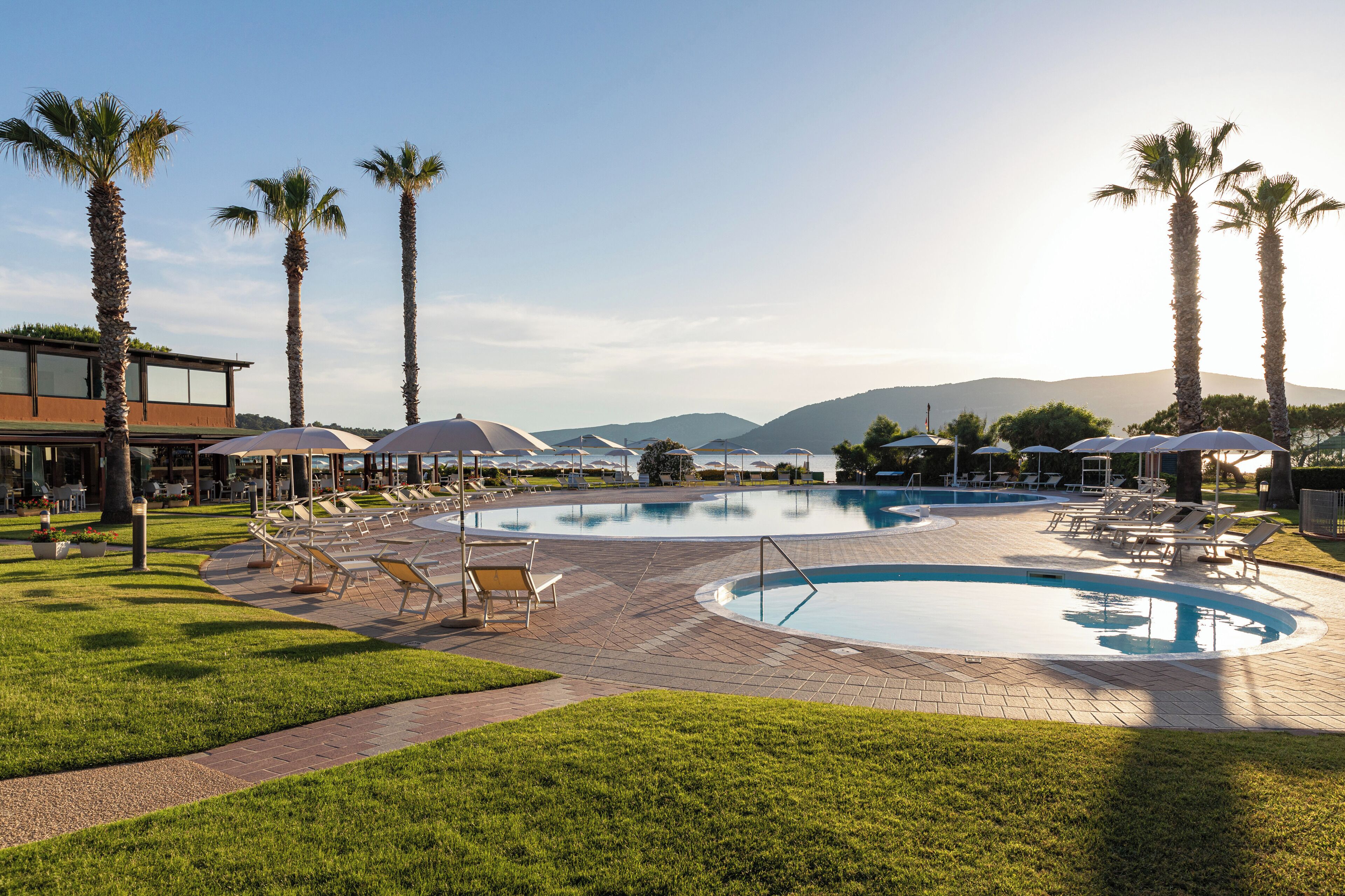 A pool area with lounge chairs and umbrellas, surrounded by palm trees and a building with large windows.