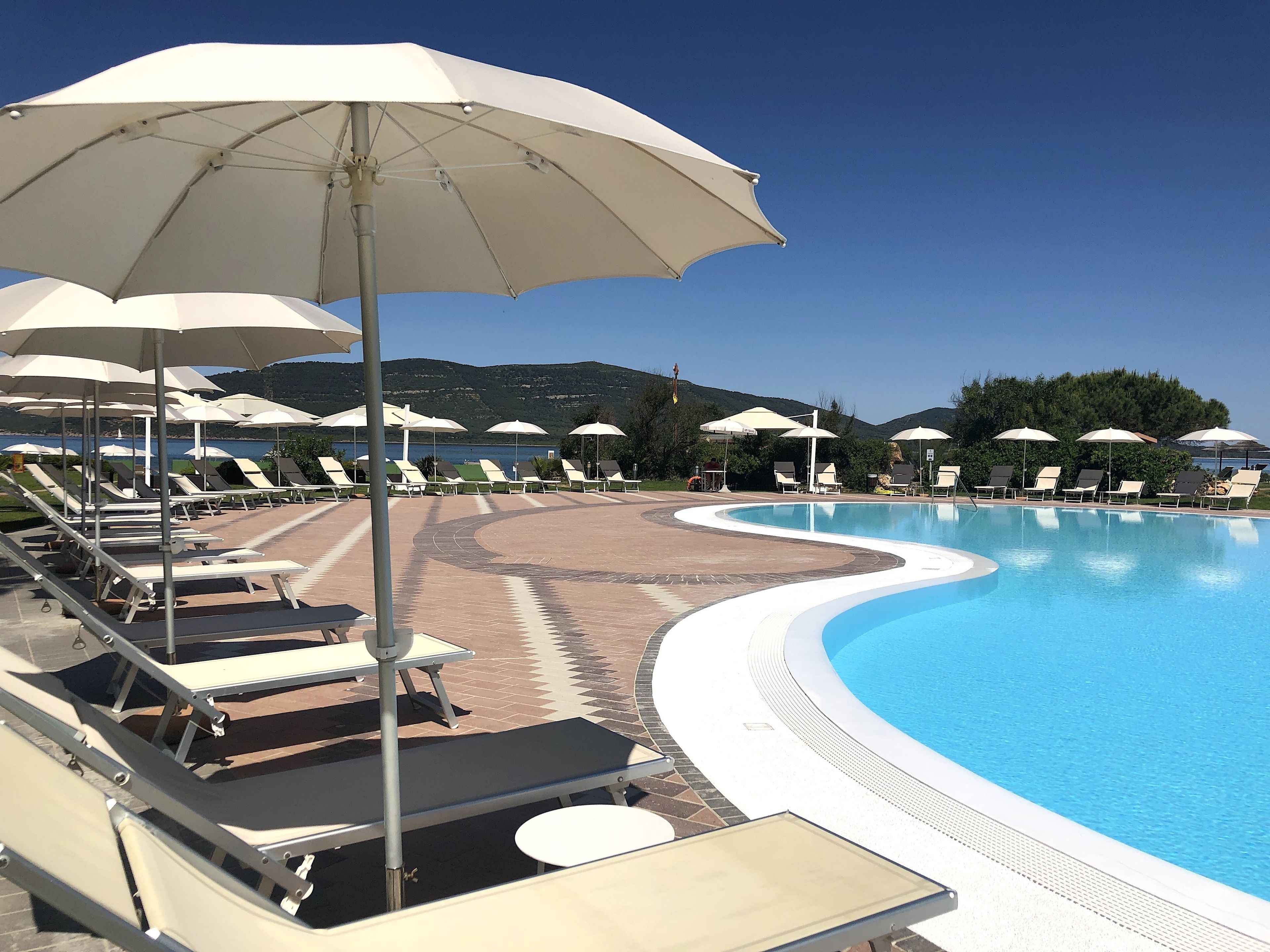 A poolside area with sun loungers and umbrellas, a clear blue pool, and a mountainous backdrop.