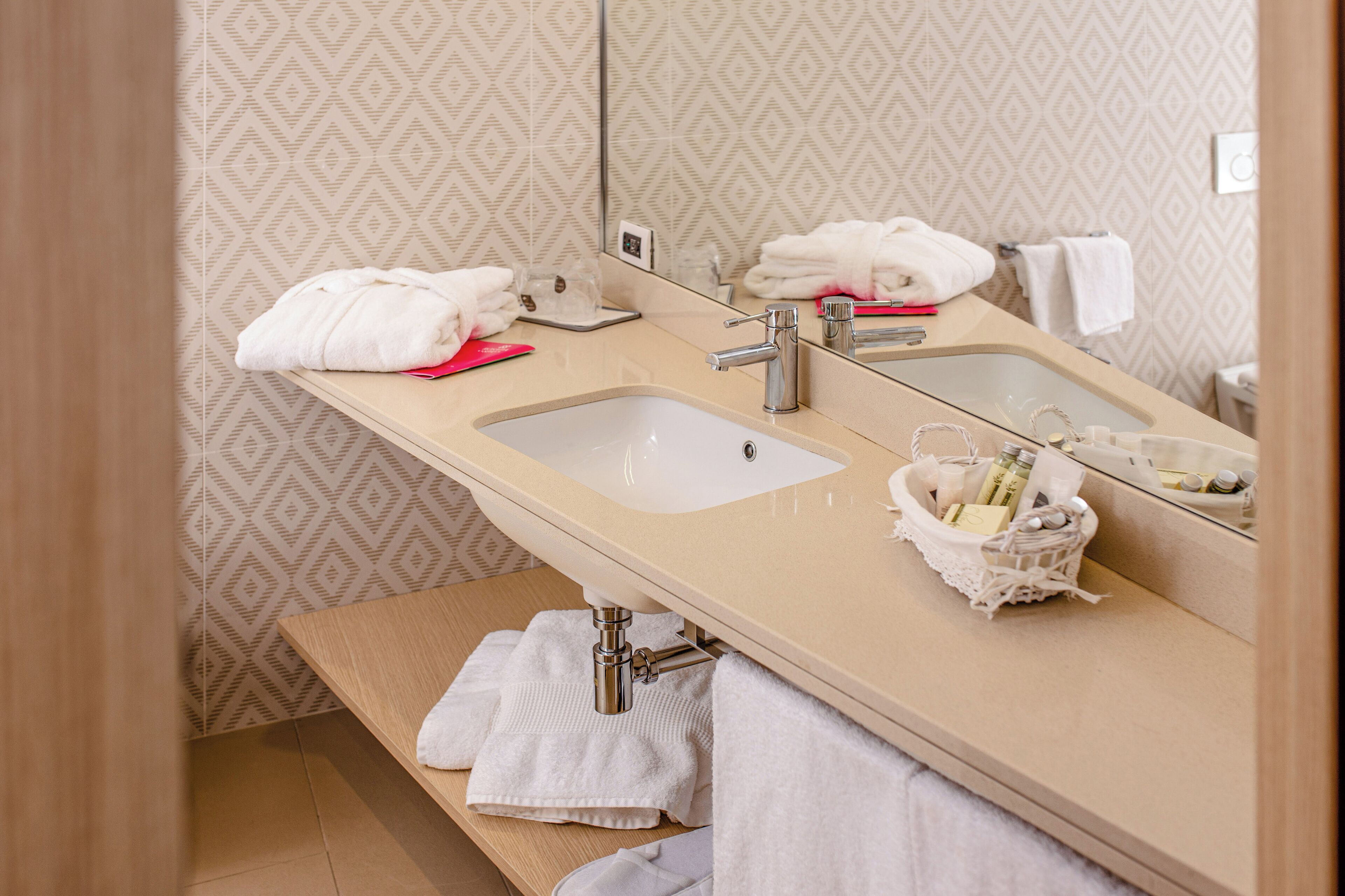 A bathroom with a beige sink, a mirror, and a towel rack. There are towels and a basket of toiletries on the counter.