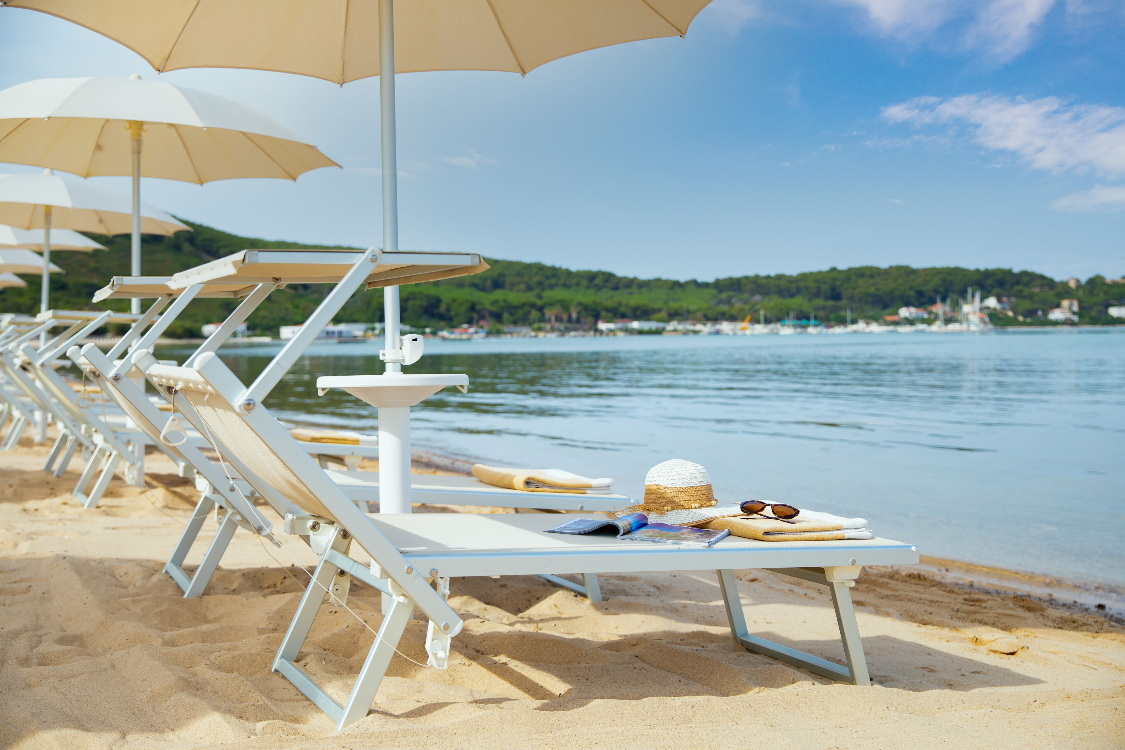 File di sedie da spiaggia bianche con ombrelloni beige, un tavolino con un cappello e occhiali da sole, e vista sul mare e su un pendio verde sullo sfondo.