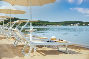 File di sedie da spiaggia bianche con ombrelloni beige, un tavolino con un cappello e occhiali da sole, e vista sul mare e su un pendio verde sullo sfondo.