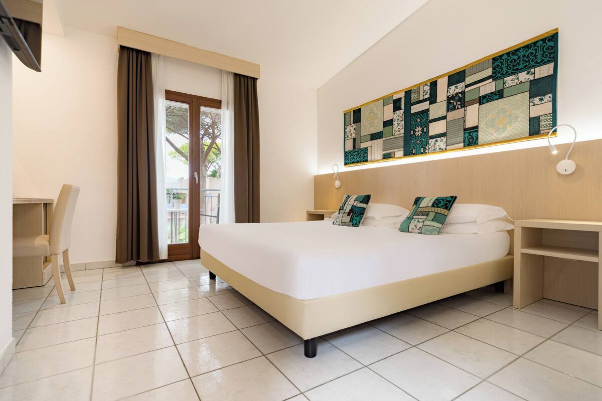 A hotel room with a large bed, a desk, a chair, a window with curtains, and a wall-mounted artwork.