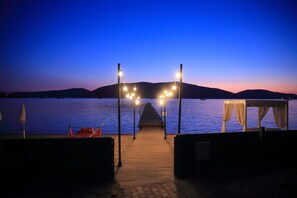 A wooden dock leading to a beach with a canopy and a table, overlooking a calm body of water and distant hills.
