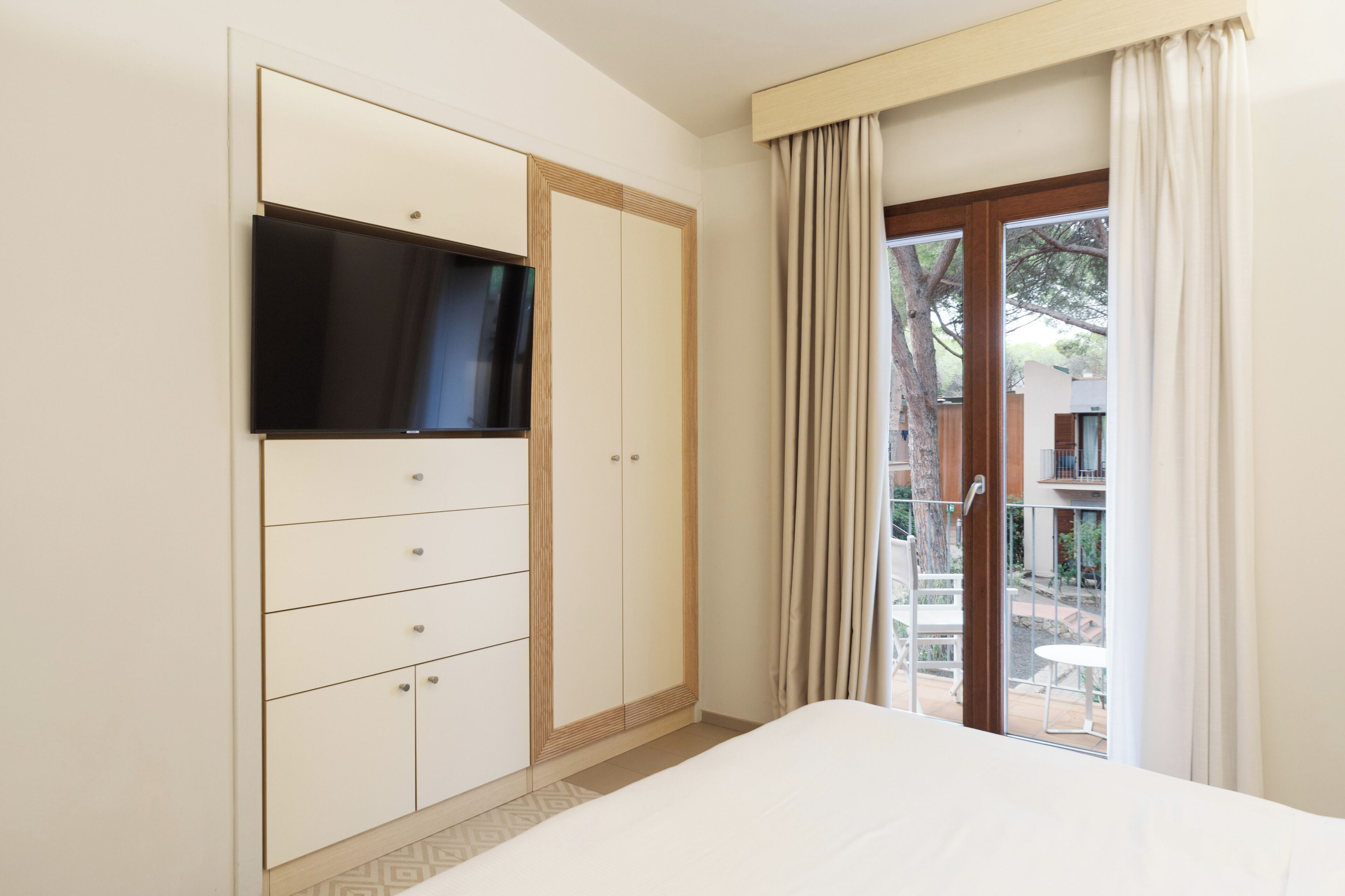 Un dormitorio con cama, armario, televisor montado en la pared y balcón con cortinas.