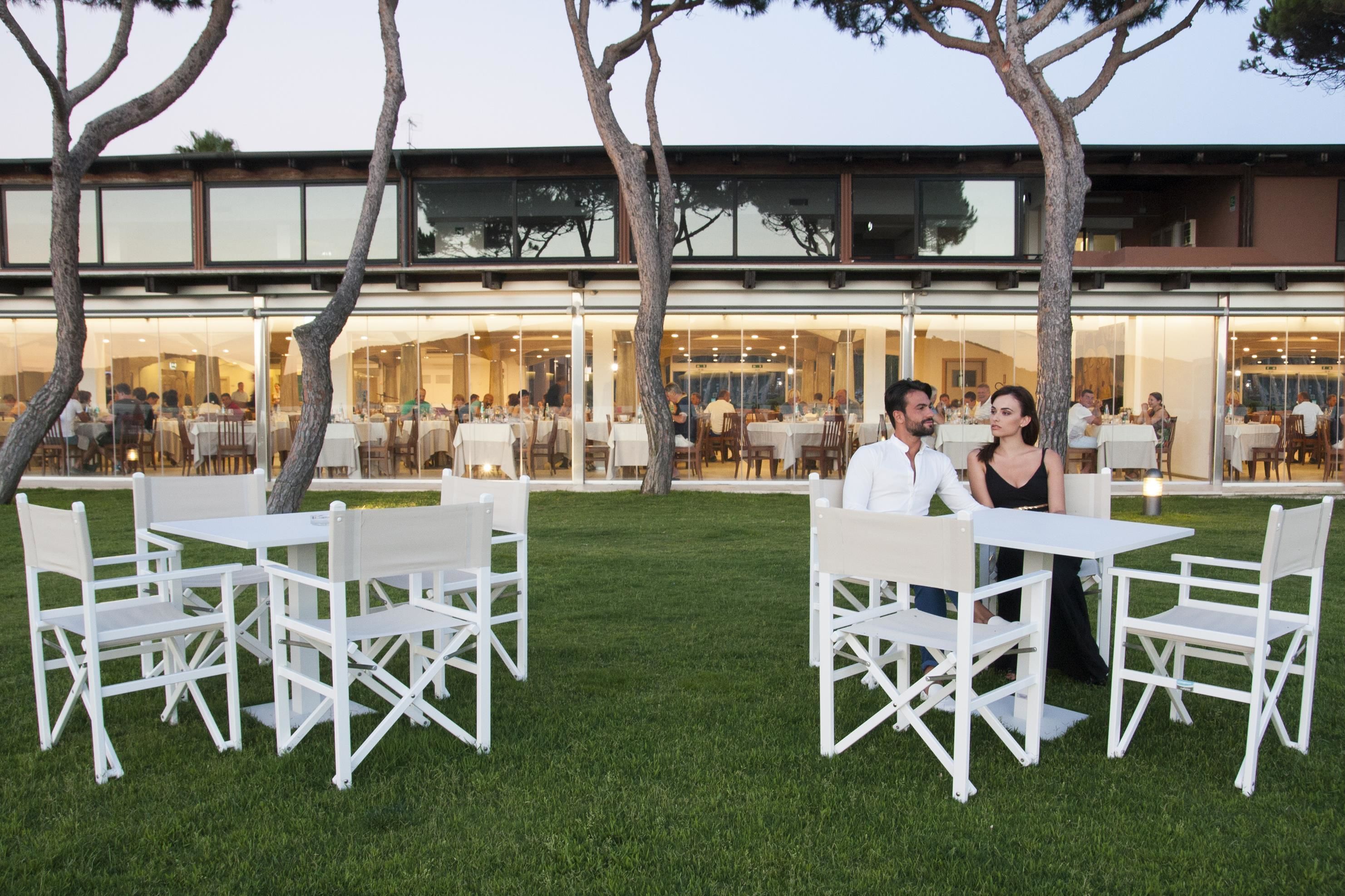 An outdoor dining area with white tables and chairs, surrounded by a well-maintained lawn and trees.