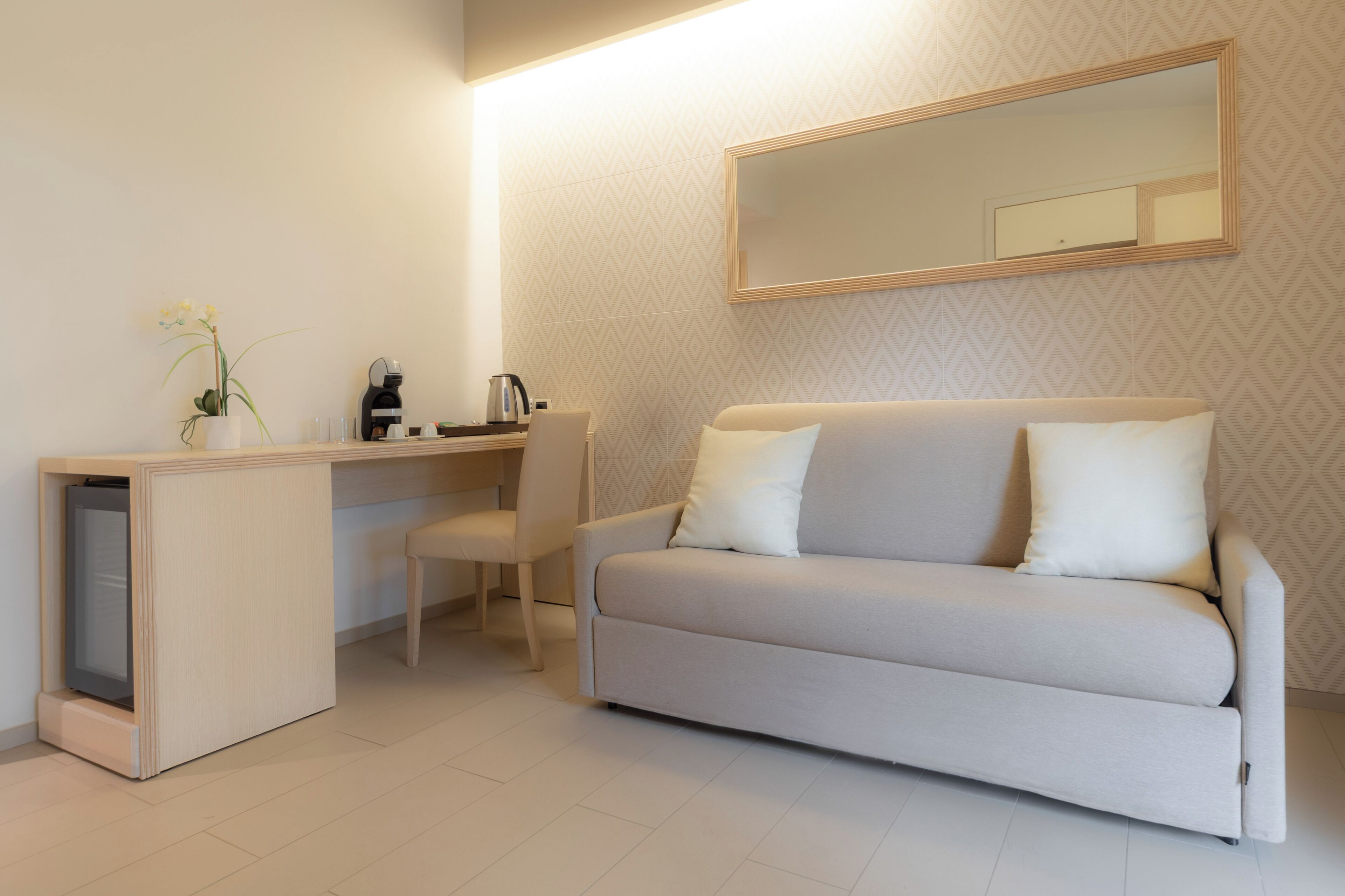 A modern room with a sofa, a desk with a chair, a coffee maker, and a vase with flowers.