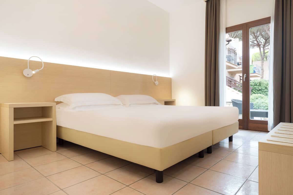 A modern hotel room with a bed, bedside tables, a wooden headboard, a view through a glass door, and a small wooden desk.