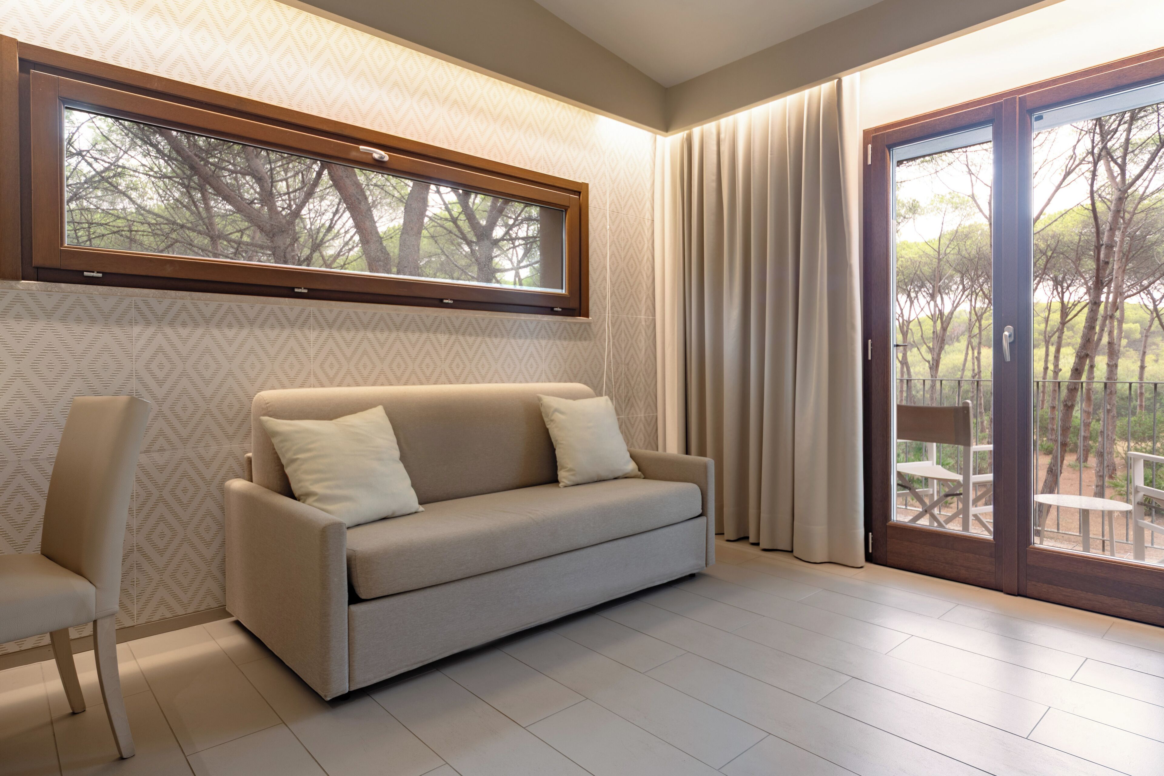 Ein modernes Wohnzimmer mit einem beigen Sofa, einem Sessel, einem Fenster mit Blick auf Bäume und einer Schiebeglastür zu einem Außenbereich.
