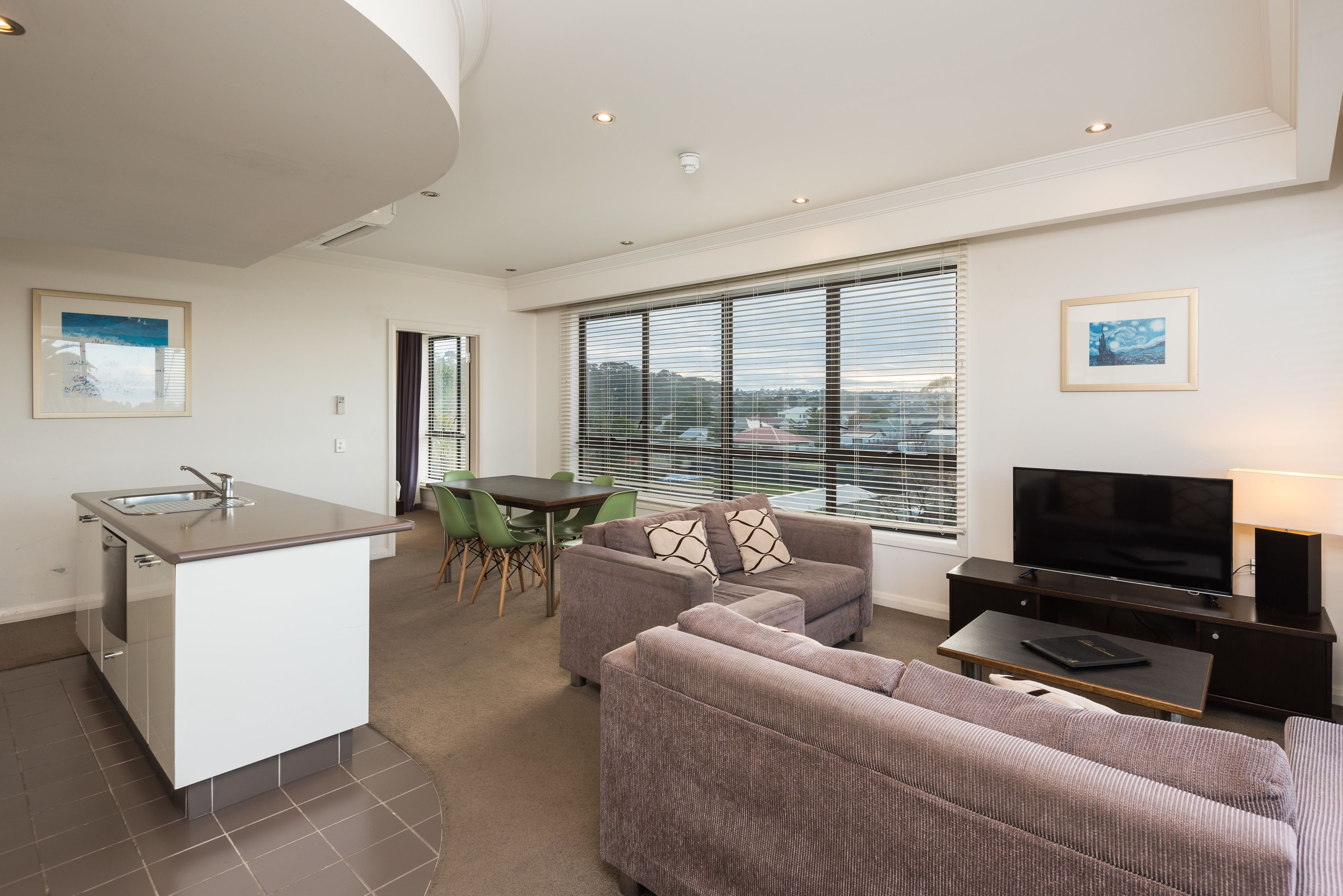 A modern apartment with a kitchenette, a dining area, a living room with a sofa and TV, and large windows with blinds.
