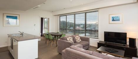 A modern apartment with a kitchenette, a dining area, a living room with a sofa and TV, and large windows with blinds.
