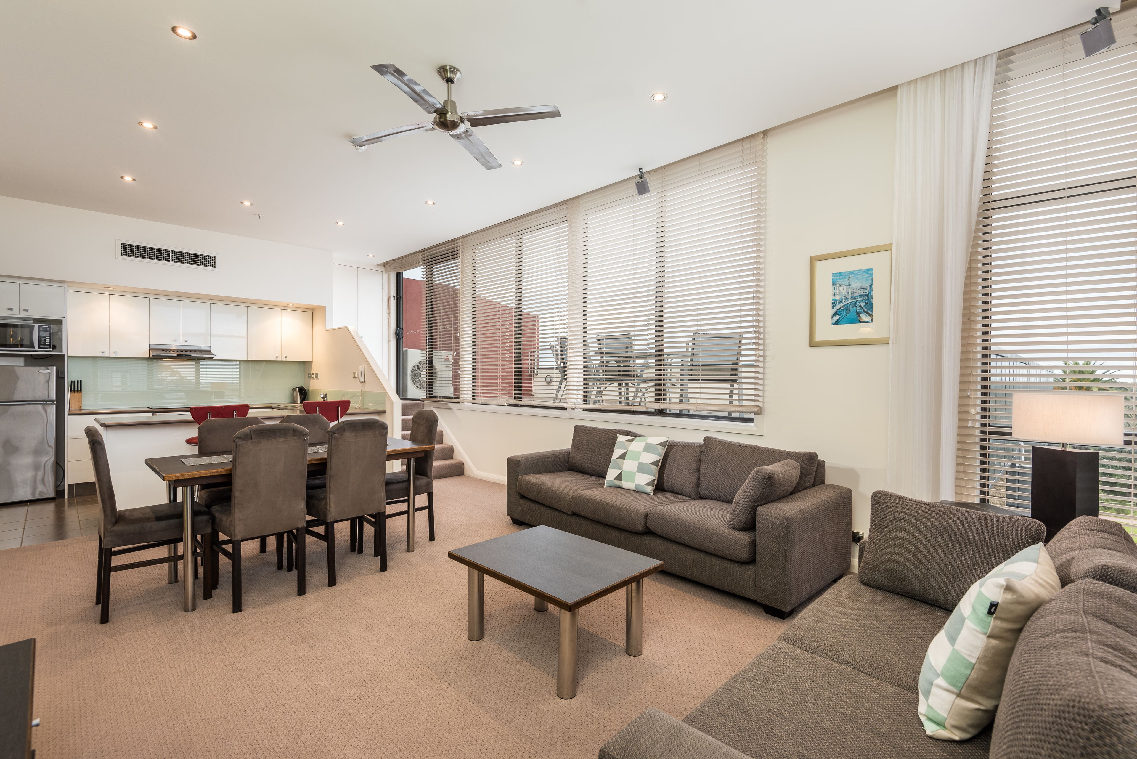 A modern living room with a sofa, coffee table, and dining area. There is a kitchen in the background with a refrigerator, microwave, and stove.