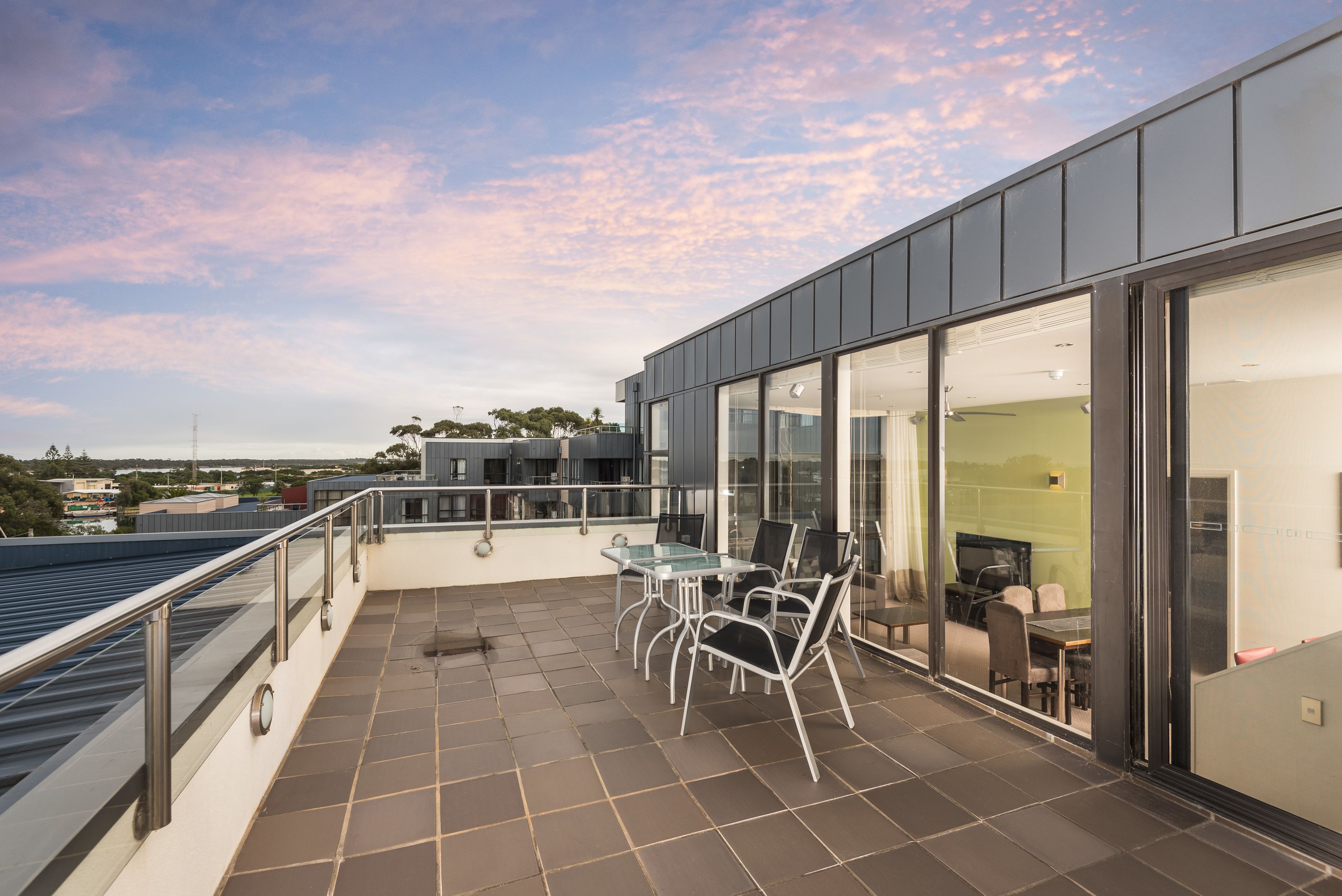 A modern outdoor terrace with a glass table and chairs, a view of the city, and a large sliding glass door leading to an interior space.