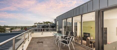 A modern outdoor terrace with a glass table and chairs, a view of the city, and a large sliding glass door leading to an interior space.