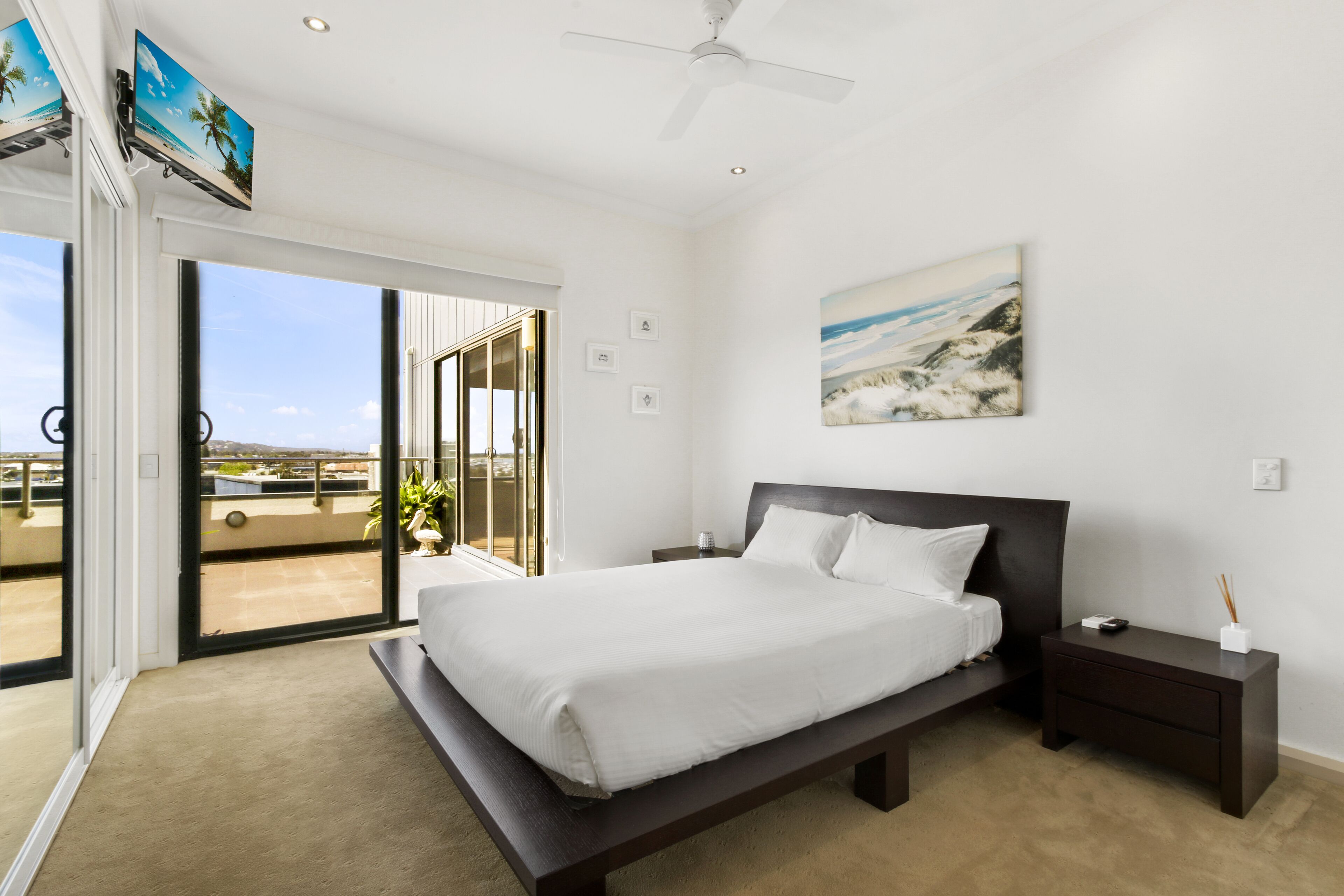 A bedroom with a large bed, a nightstand, a ceiling fan, a painting, and a view through a sliding glass door to an outdoor balcony.